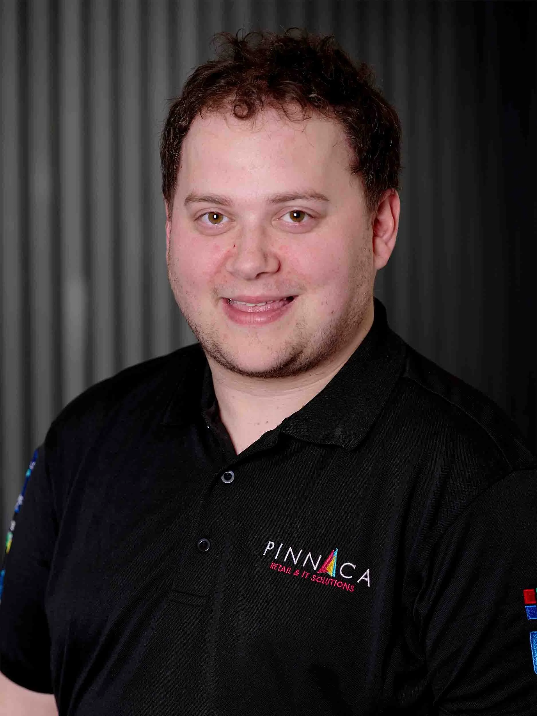 A young man with short, curly brown hair and light skin smiling, wearing a black polo shirt with the logos 'PINNICA Retail & IT Solutions' on the chest and sleeves, against a dark, vertically striped background.