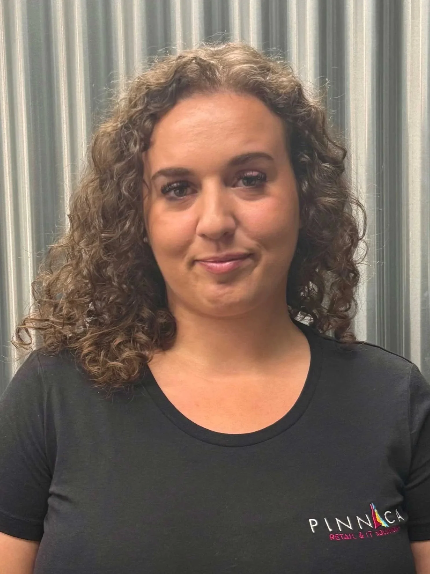 A woman with curly brown hair, wearing a black t-shirt with 'Pinnaca' logo, standing in front of a metallic, ribbed background.