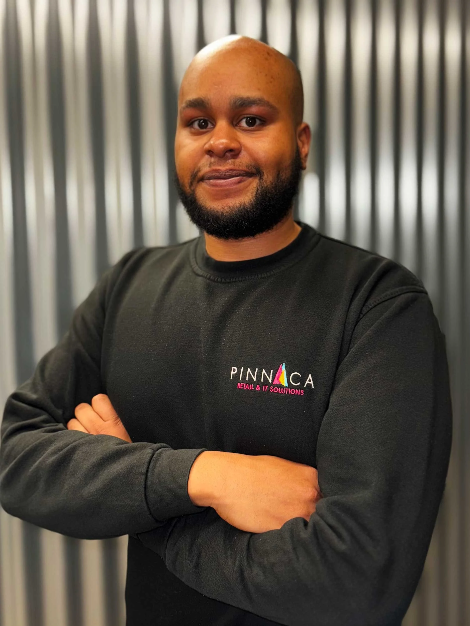 A man with a bald head and beard wearing a black sweatshirt with 'PINNACA RETAIL & IT SOLUTIONS' logo, standing with arms crossed, in front of a metallic striped background.