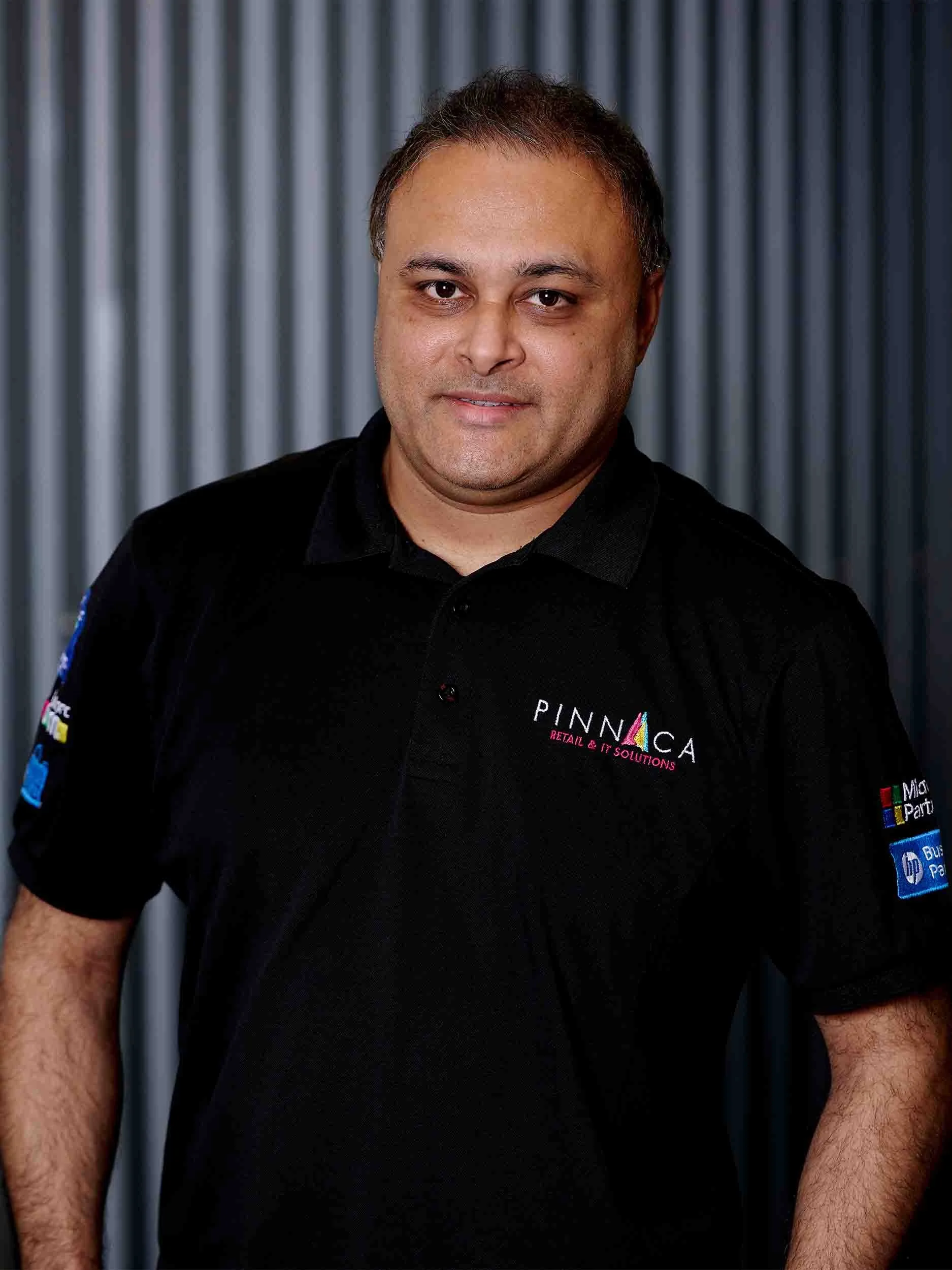 A man wearing a black polo shirt with logos stands in front of a metallic background.