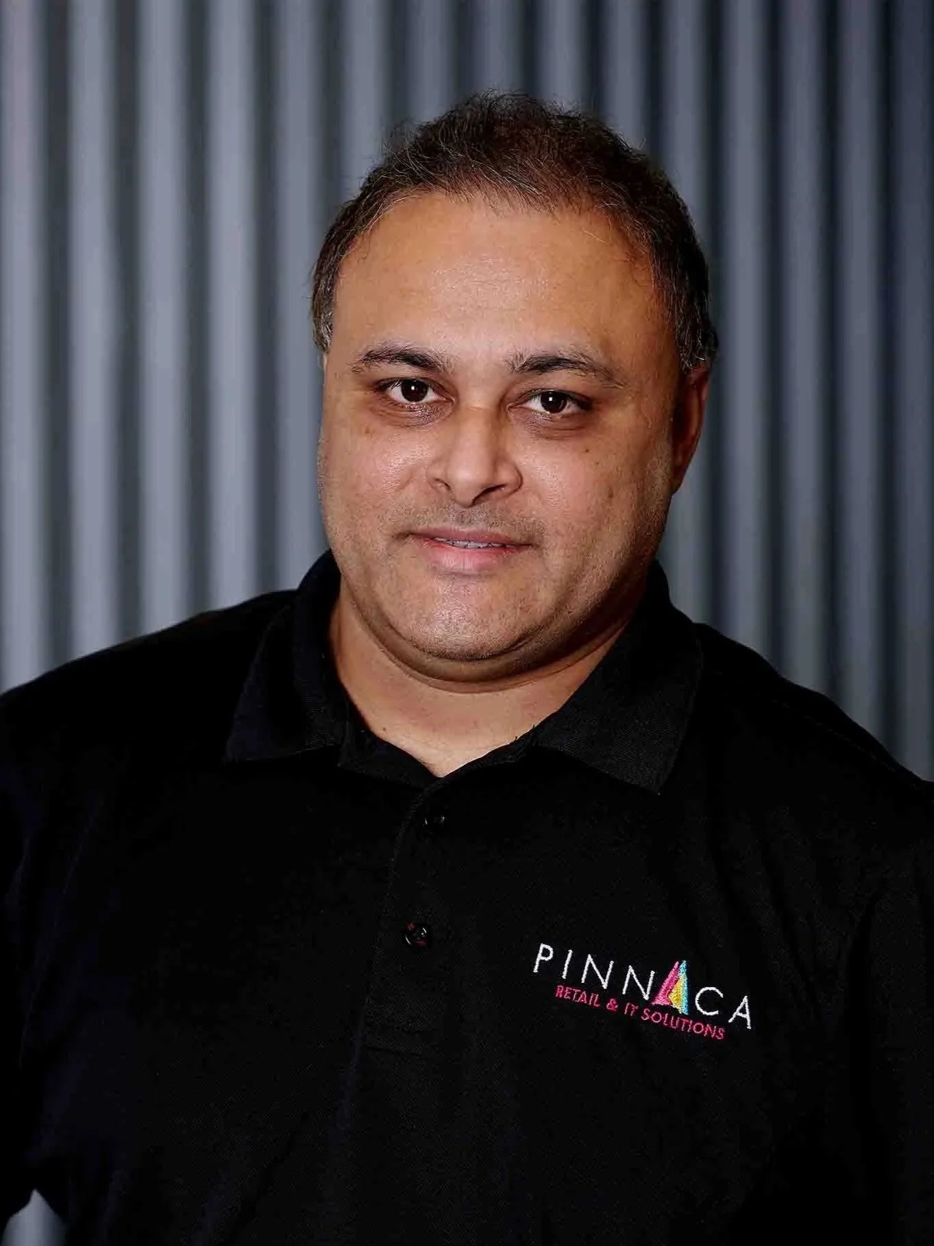 A man wearing a black polo shirt with logos stands in front of a metallic background.