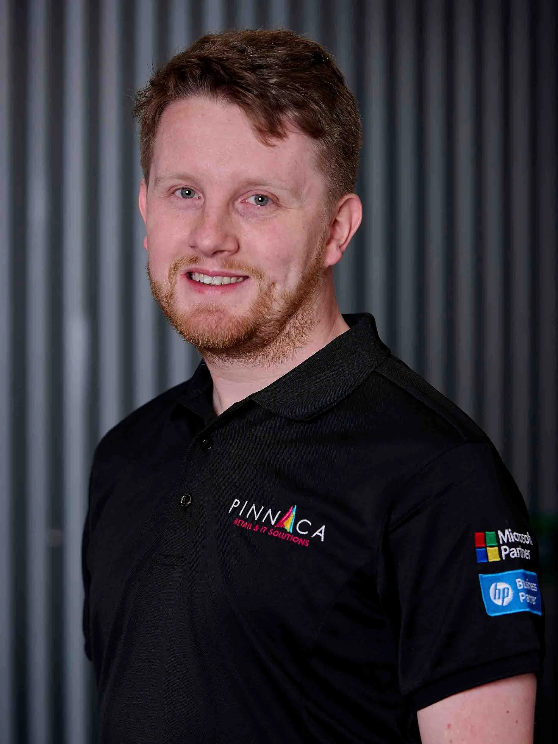 A man with red hair and a beard smiling, wearing a black polo shirt with logos for Pinnaca, Microsoft Partner, and HP, standing in front of a background with vertical gray and black stripes.