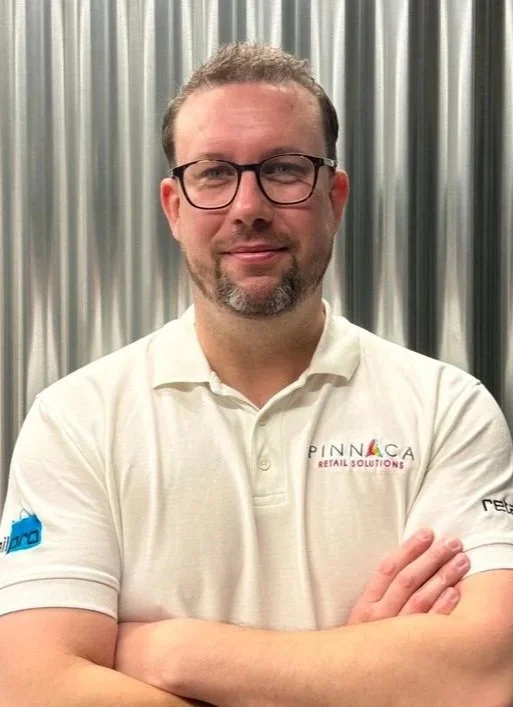 A smiling man with glasses and short brown hair, wearing a black polo shirt with a Pinnaca logo, standing in front of a staircase.