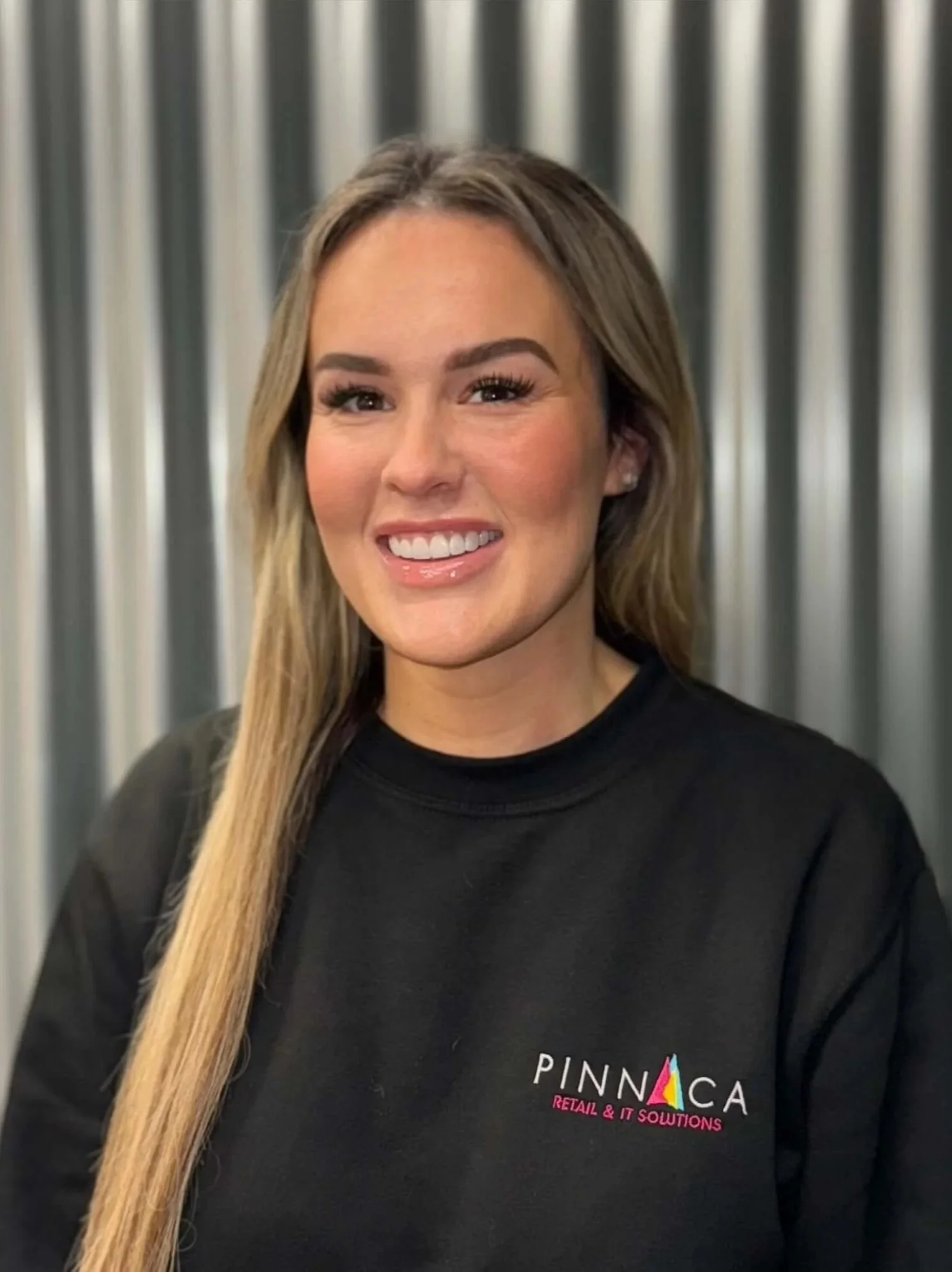A woman smiling, wearing a black sweatshirt with the logo "PINNACA Retail & IT Solutions" and standing in front of a metallic, corrugated background.