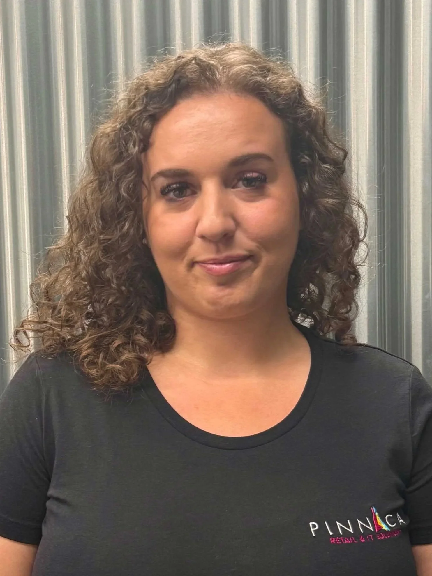 A woman with curly brown hair, wearing a black t-shirt with 'Pinnaca' logo, standing in front of a metallic, ribbed background.