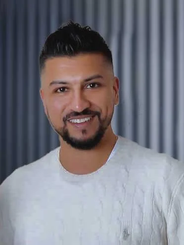 A man with short dark hair, a beard, and mustache smiling at the camera, wearing a light-colored sweater with a vertical striped curtain background.