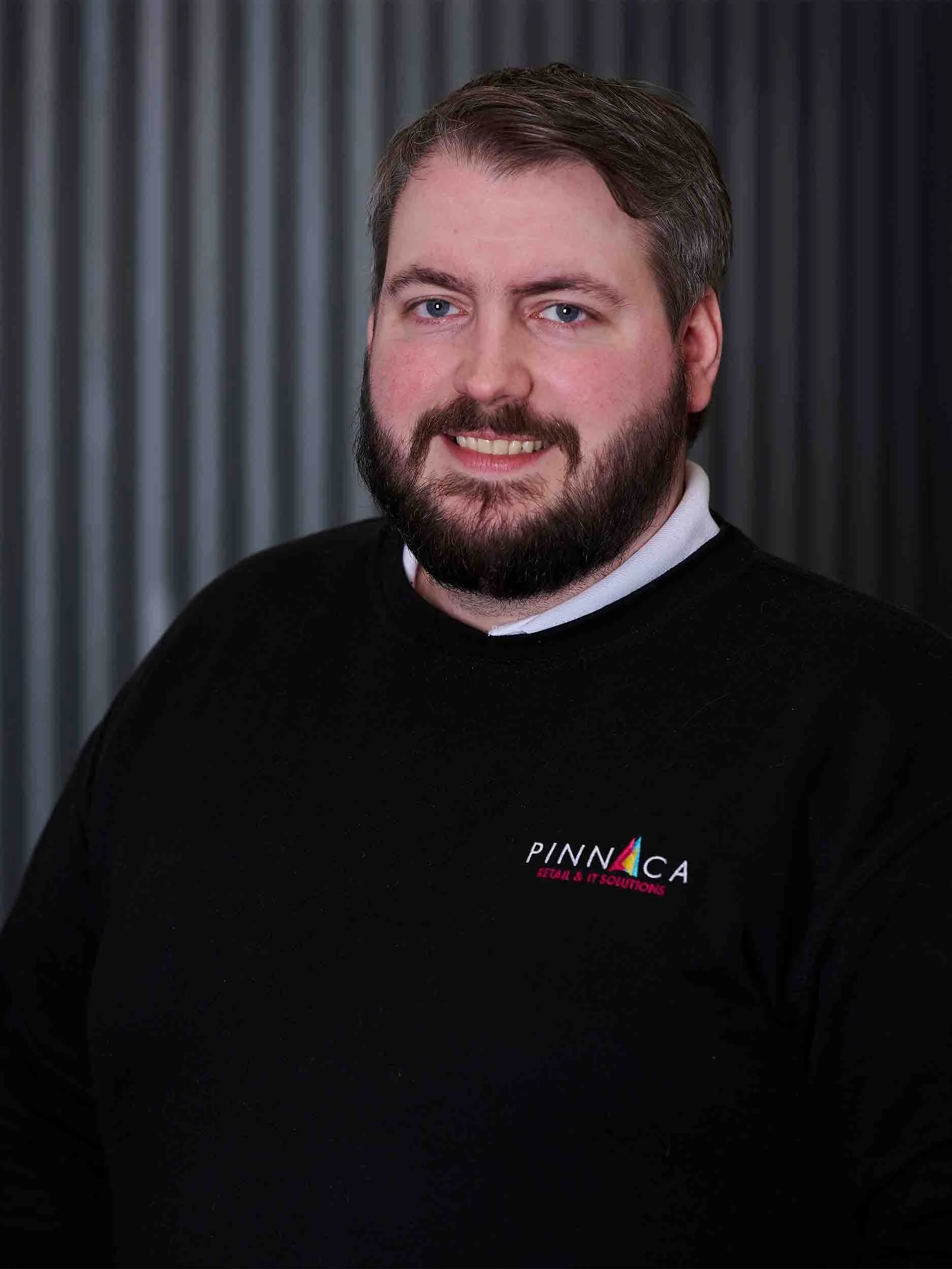 A man with a beard and light blue eyes, wearing a black sweater with a colorful logo and the words "PINNACA RETAIL & IT SOLUTIONS" embroidered on it, smiling in front of a striped gray background.