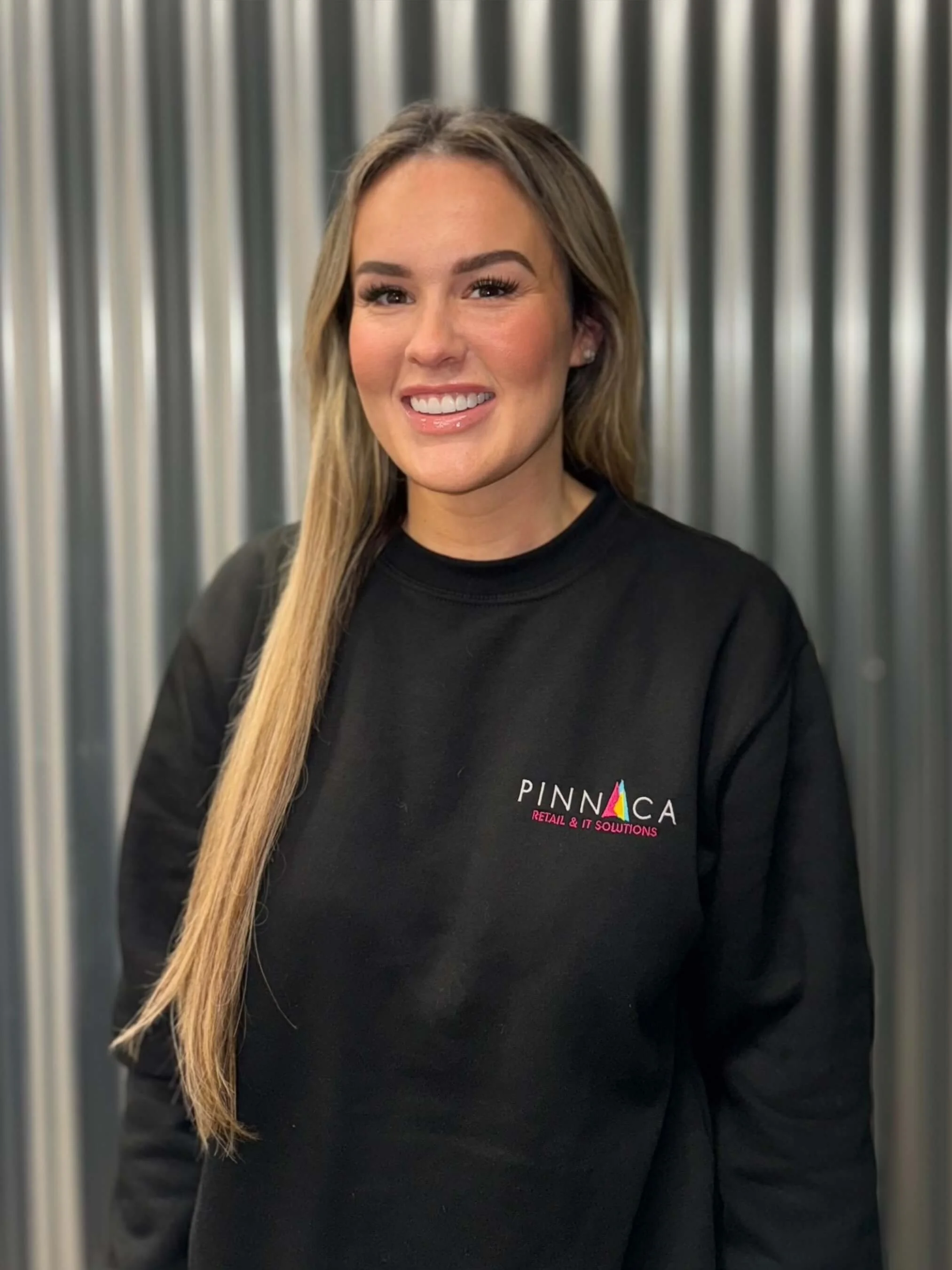 A woman smiling, wearing a black sweatshirt with the logo "PINNACA Retail & IT Solutions" and standing in front of a metallic, corrugated background.