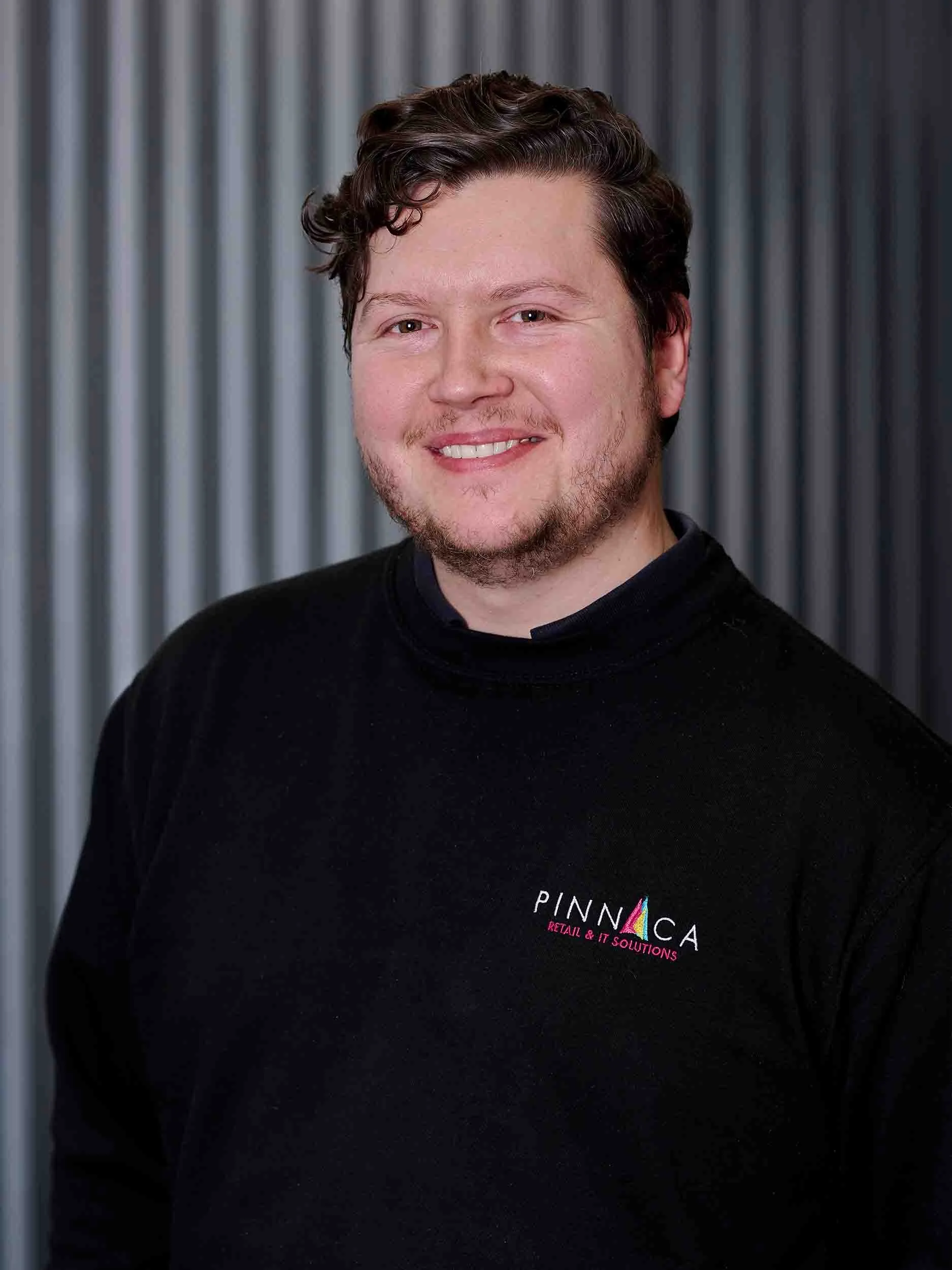 A man with curly brown hair and a beard, smiling, wearing a black shirt with the logo "PINNICA RETAIL & IT SOLUTIONS" in front of a gray, metal-panel background.