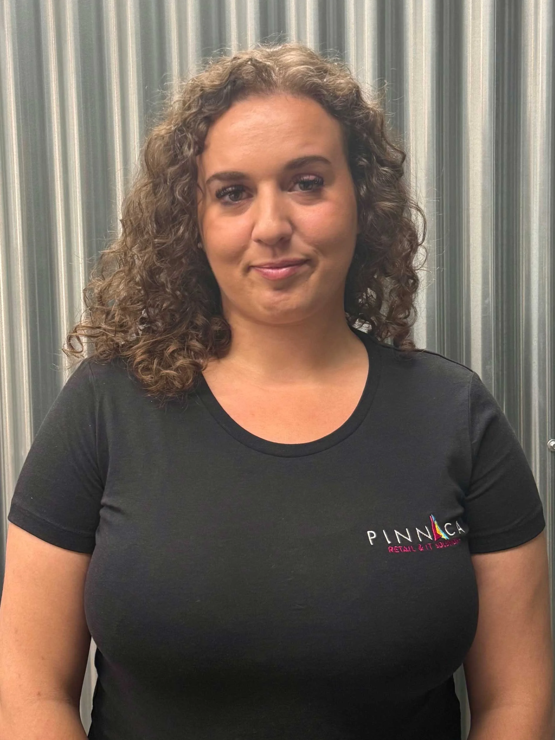 A woman with curly brown hair, wearing a black t-shirt with 'Pinnaca' logo, standing in front of a metallic, ribbed background.
