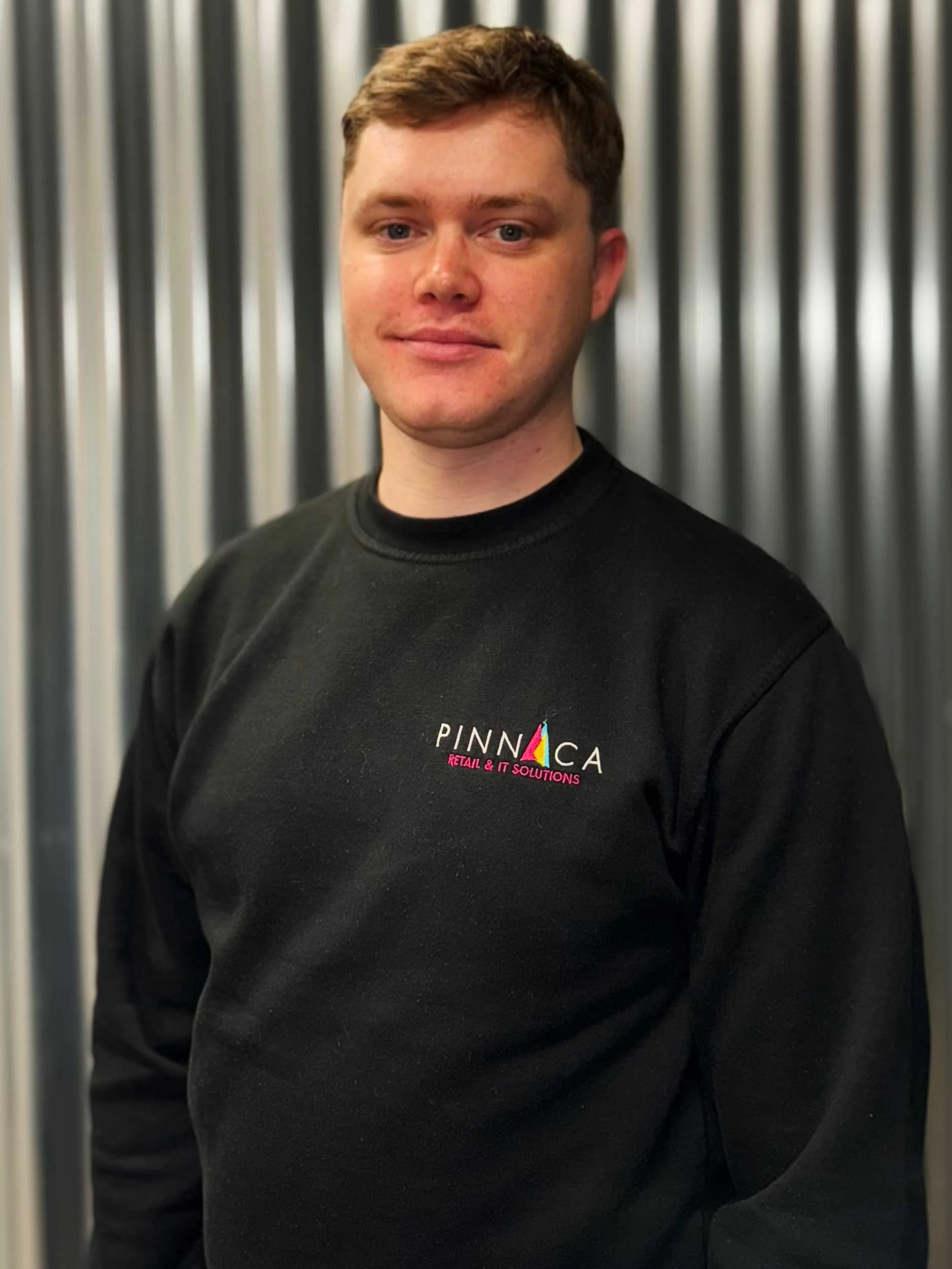 A young man with short light brown hair, wearing a black sweatshirt with a logo that reads 'PINNACA Retail & IT Solutions,' standing in front of a metallic, corrugated background.