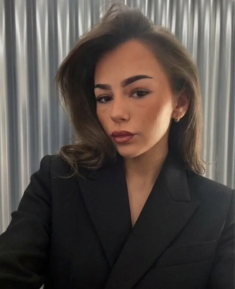 Young woman with long brown hair wearing a sleeveless black top, standing in front of a metallic curtain.