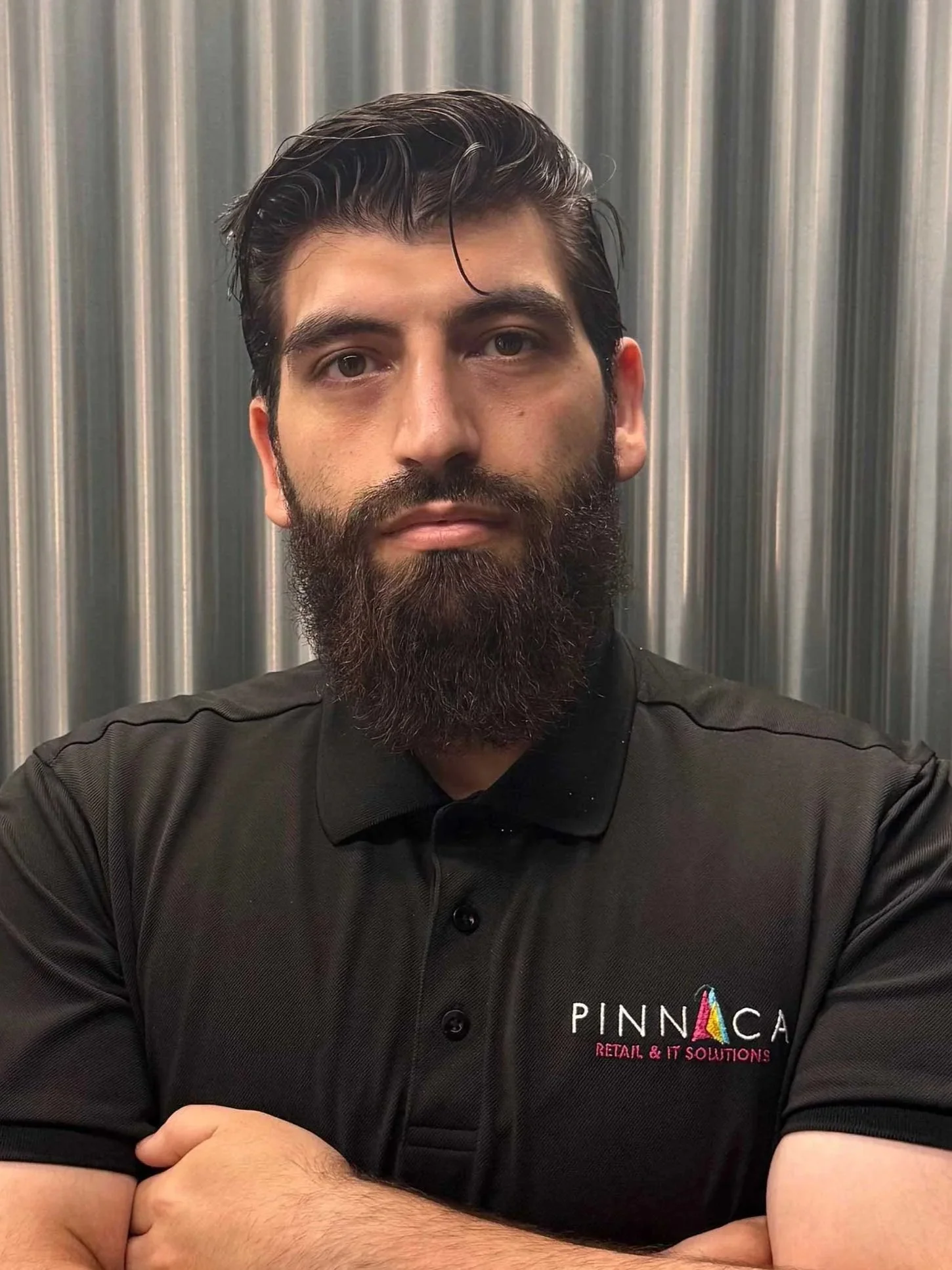 A man with dark hair and a full beard standing with arms crossed in front of a metallic background, wearing a black polo shirt with a Pinnaca logo.