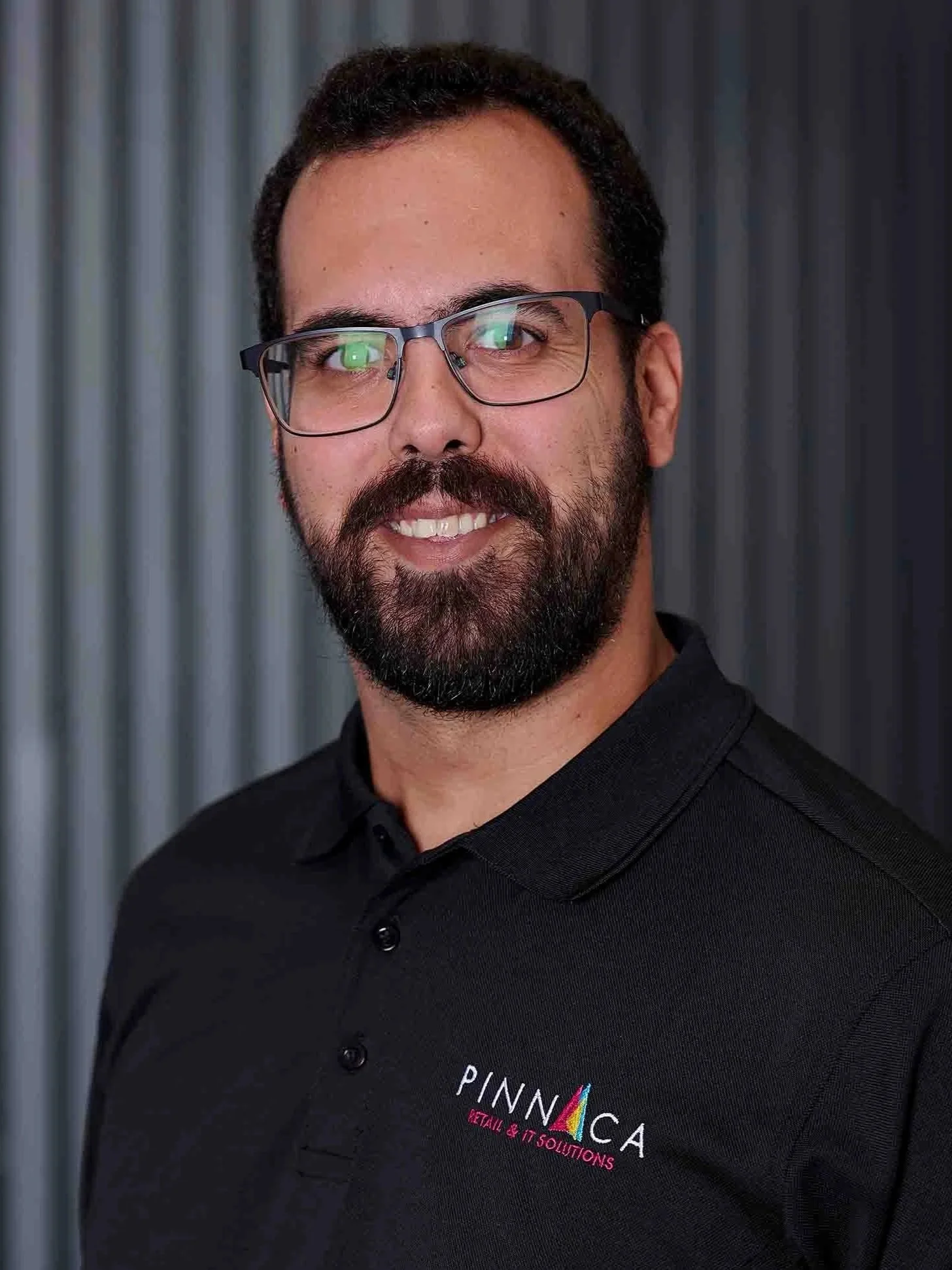 A man with glasses, a beard, and dark hair, wearing a black polo shirt with logos for Pinnaca and Microsoft, standing in front of a gray metallic background.