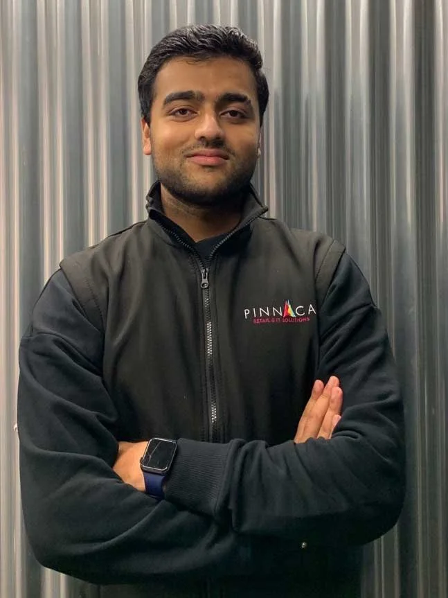 A man with dark hair standing with arms crossed in front of a metallic background, wearing a black polo shirt with a Pinnaca logo.