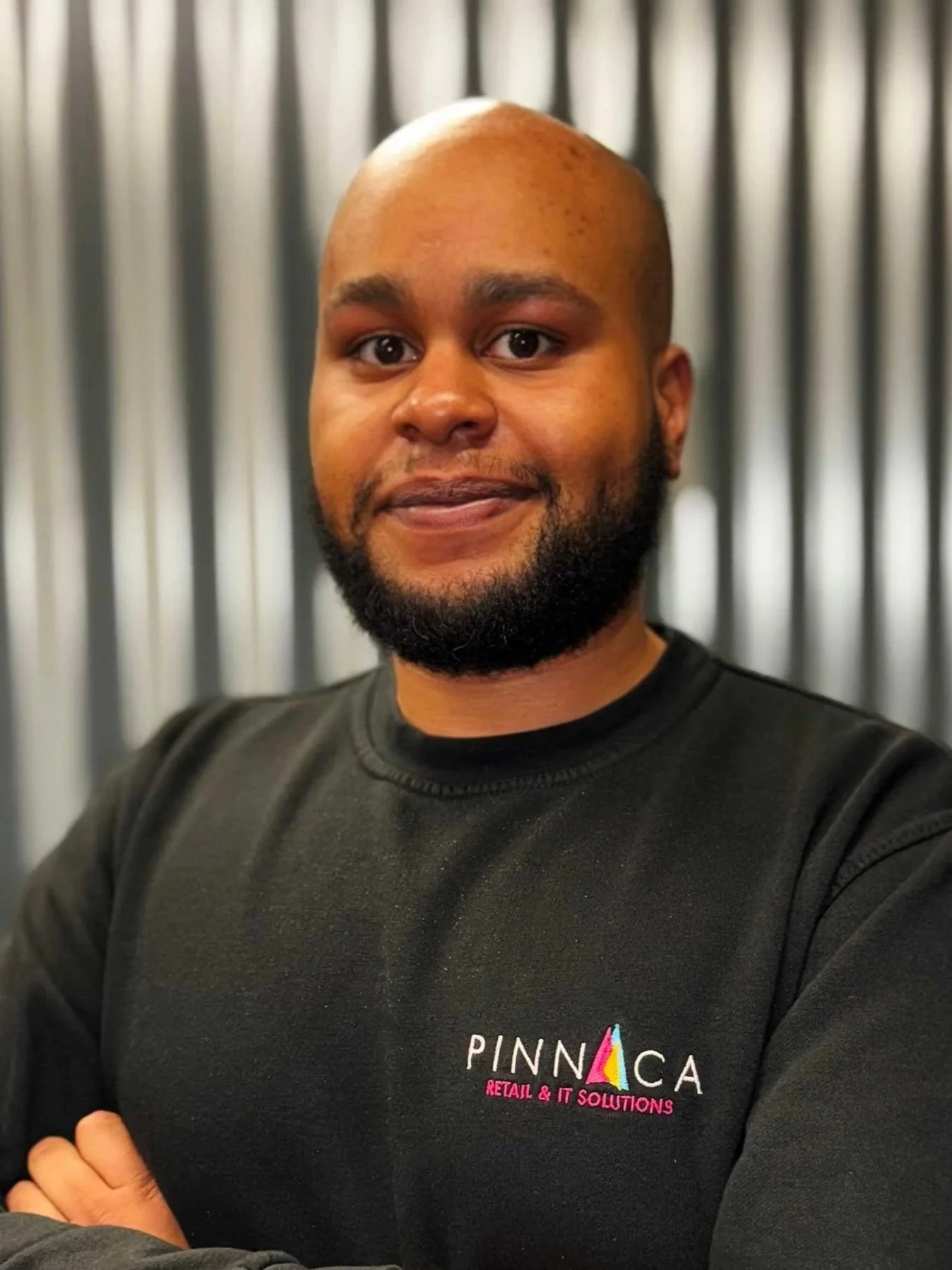 A man with a bald head and beard wearing a black sweatshirt with 'PINNACA RETAIL & IT SOLUTIONS' logo, standing with arms crossed, in front of a metallic striped background.