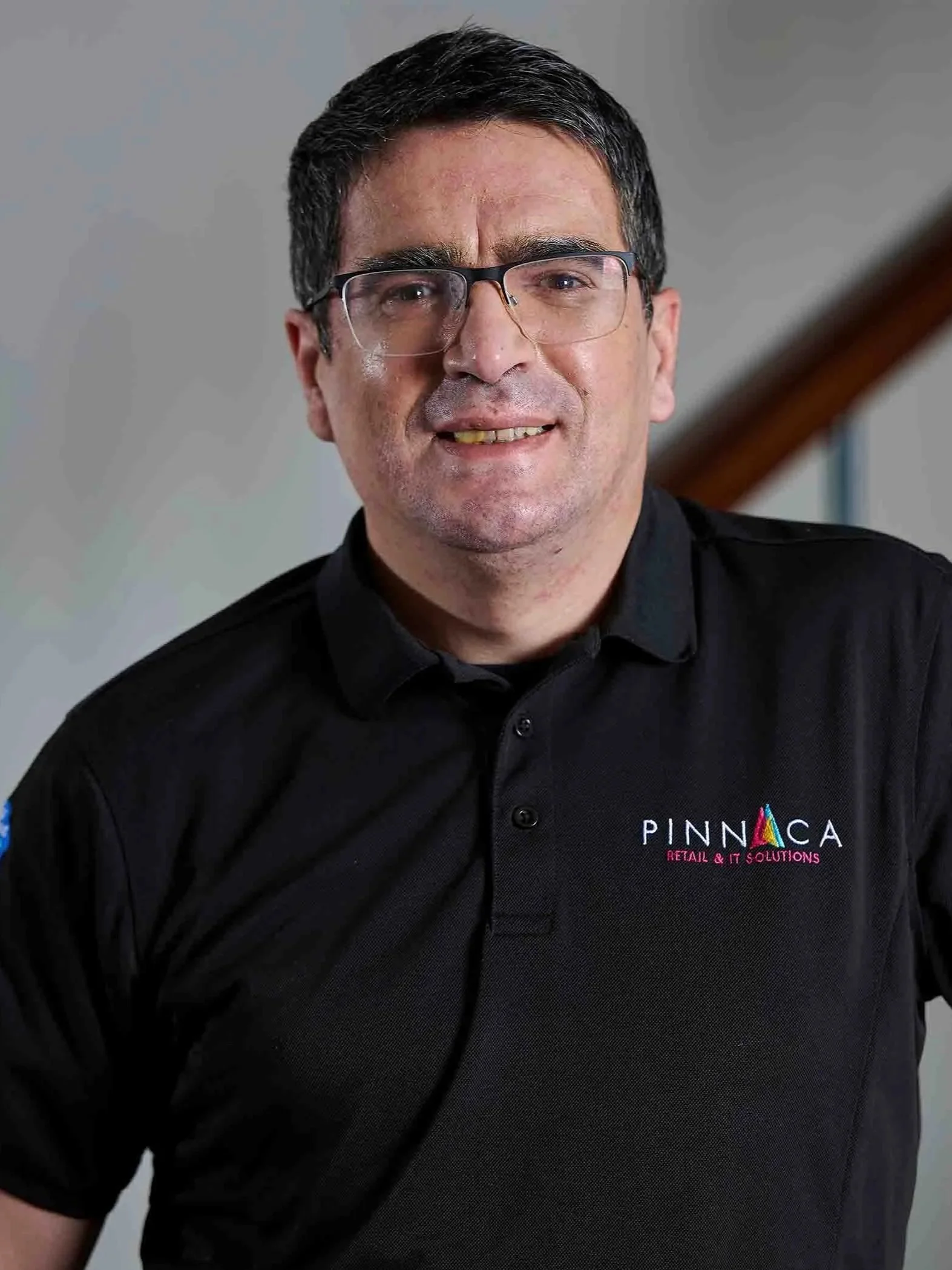 A man with glasses and short dark hair wearing a black polo shirt with a logo that says Pinnaca Retail & IT Solutions, standing indoors near a staircase.