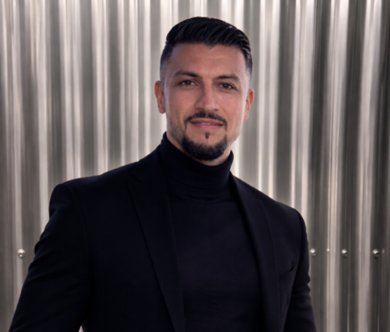 A man with short dark hair, a beard, and mustache smiling at the camera, wearing a light-colored sweater with a vertical striped curtain background.