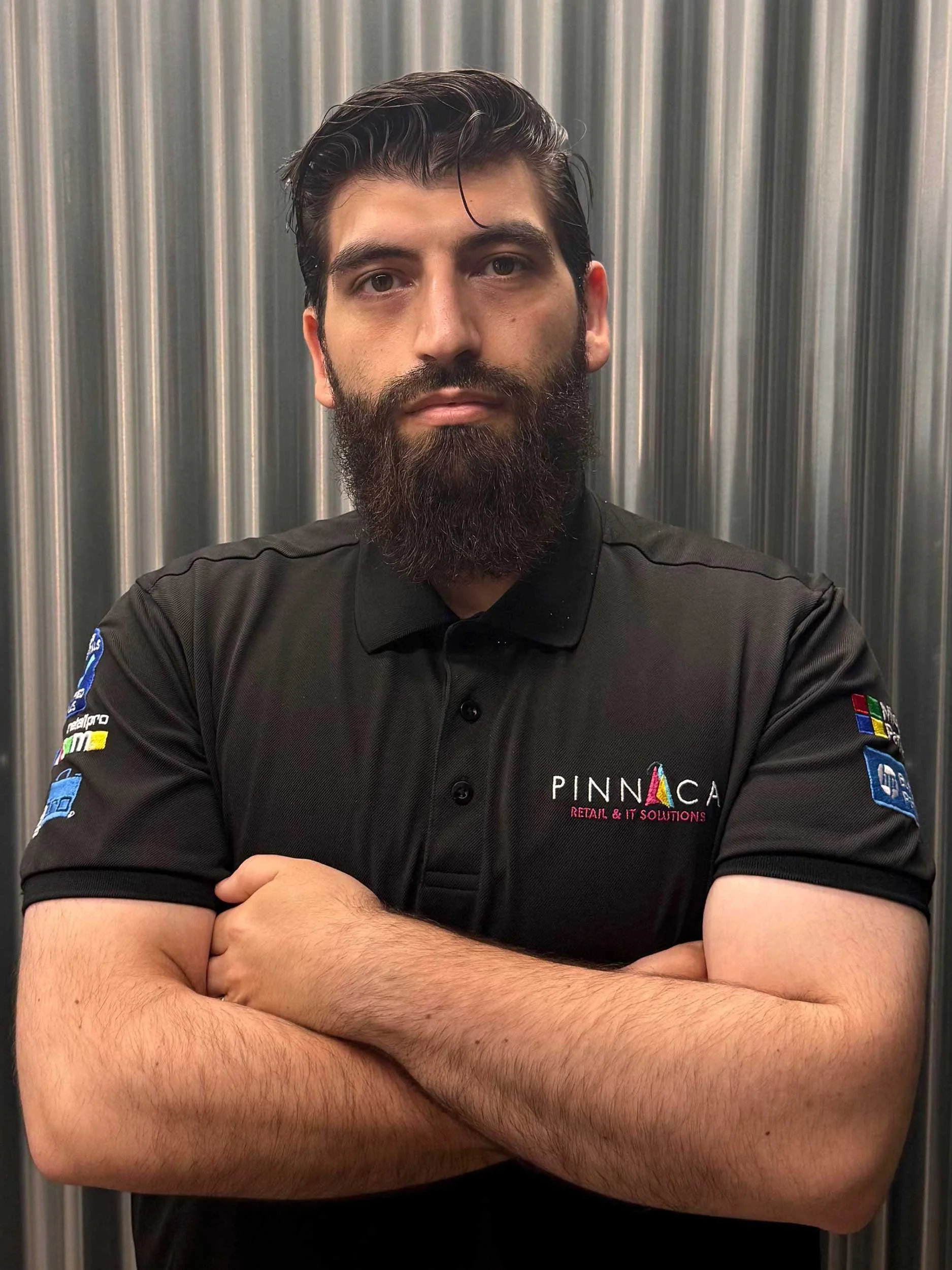 A man with dark hair and a full beard standing with arms crossed in front of a metallic background, wearing a black polo shirt with a Pinnaca logo.