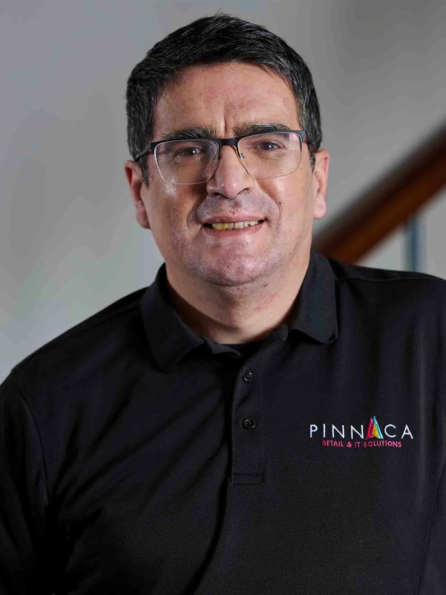 A man with glasses and short dark hair wearing a black polo shirt with a logo that says Pinnaca Retail & IT Solutions, standing indoors near a staircase.