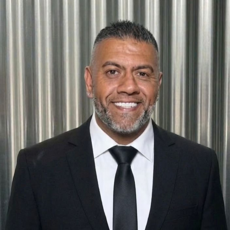 A smiling man with short hair and tattoos on his arms, wearing a black polo shirt with a logo that says Pinnaca Retail & IT Solutions, standing indoors near a staircase.