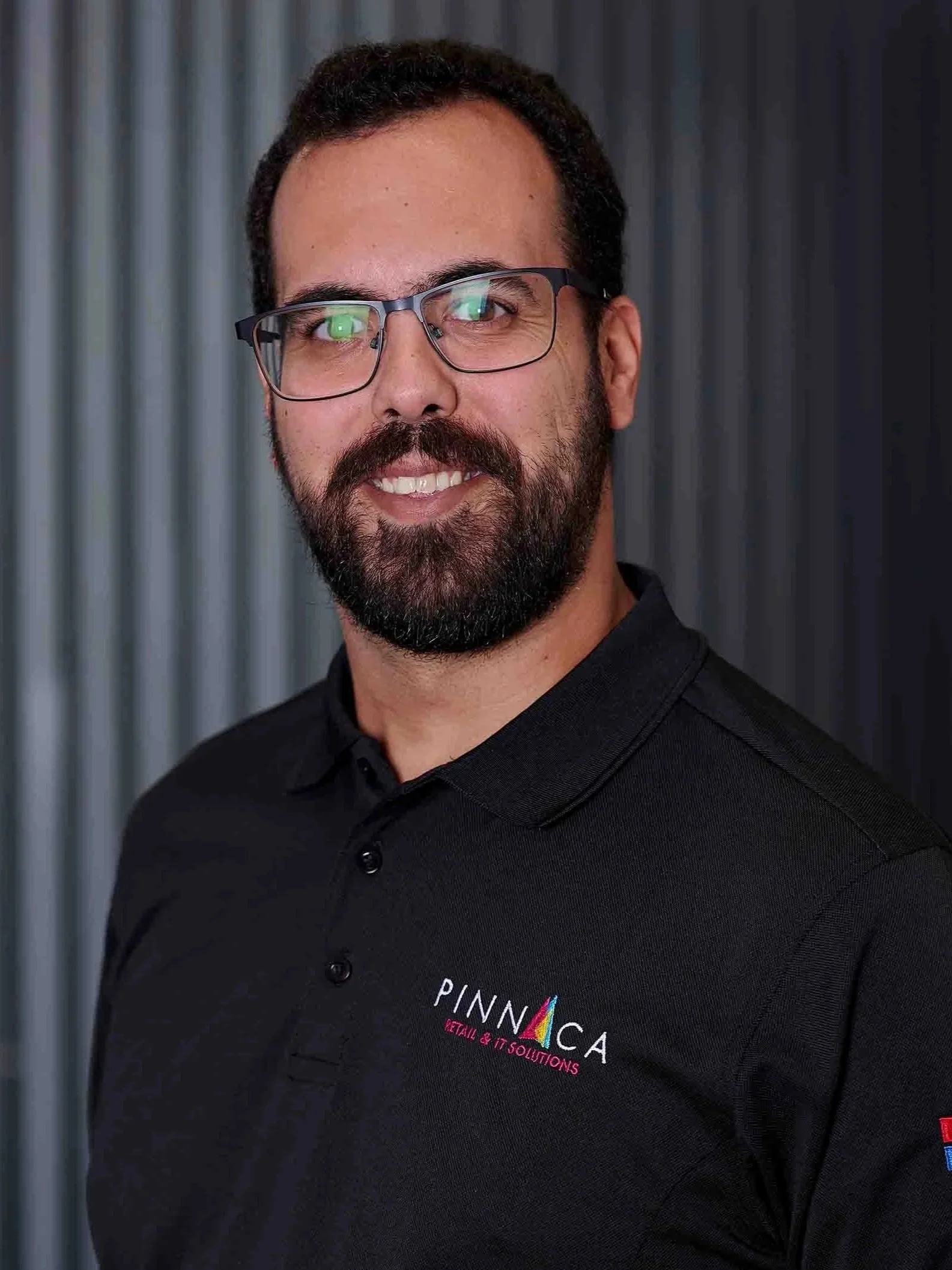 A man with glasses, a beard, and dark hair, wearing a black polo shirt with logos for Pinnaca and Microsoft, standing in front of a gray metallic background.