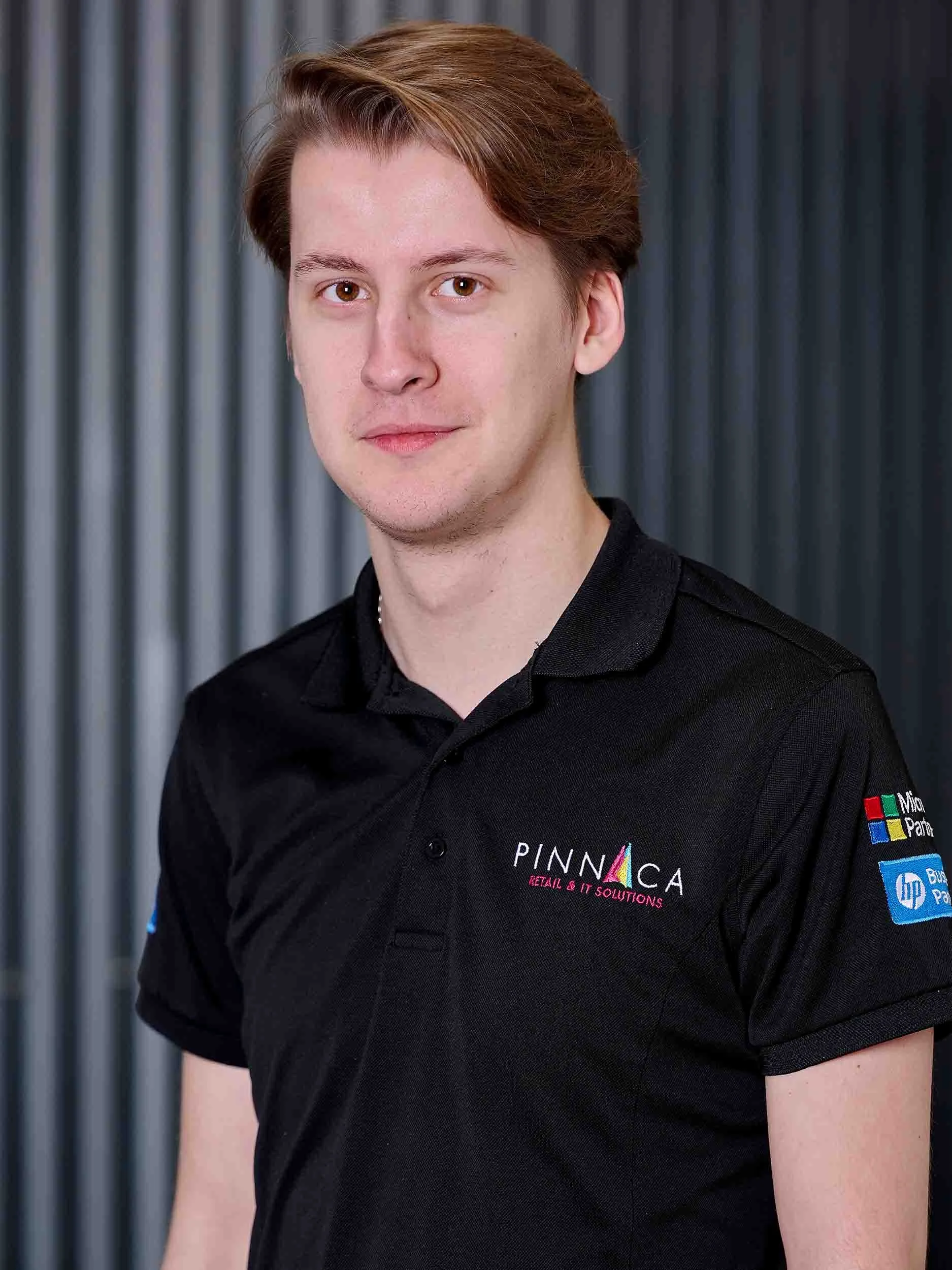 Young man with light skin, light brown hair, wearing a black polo shirt with logos, standing in front of a grey striped background.