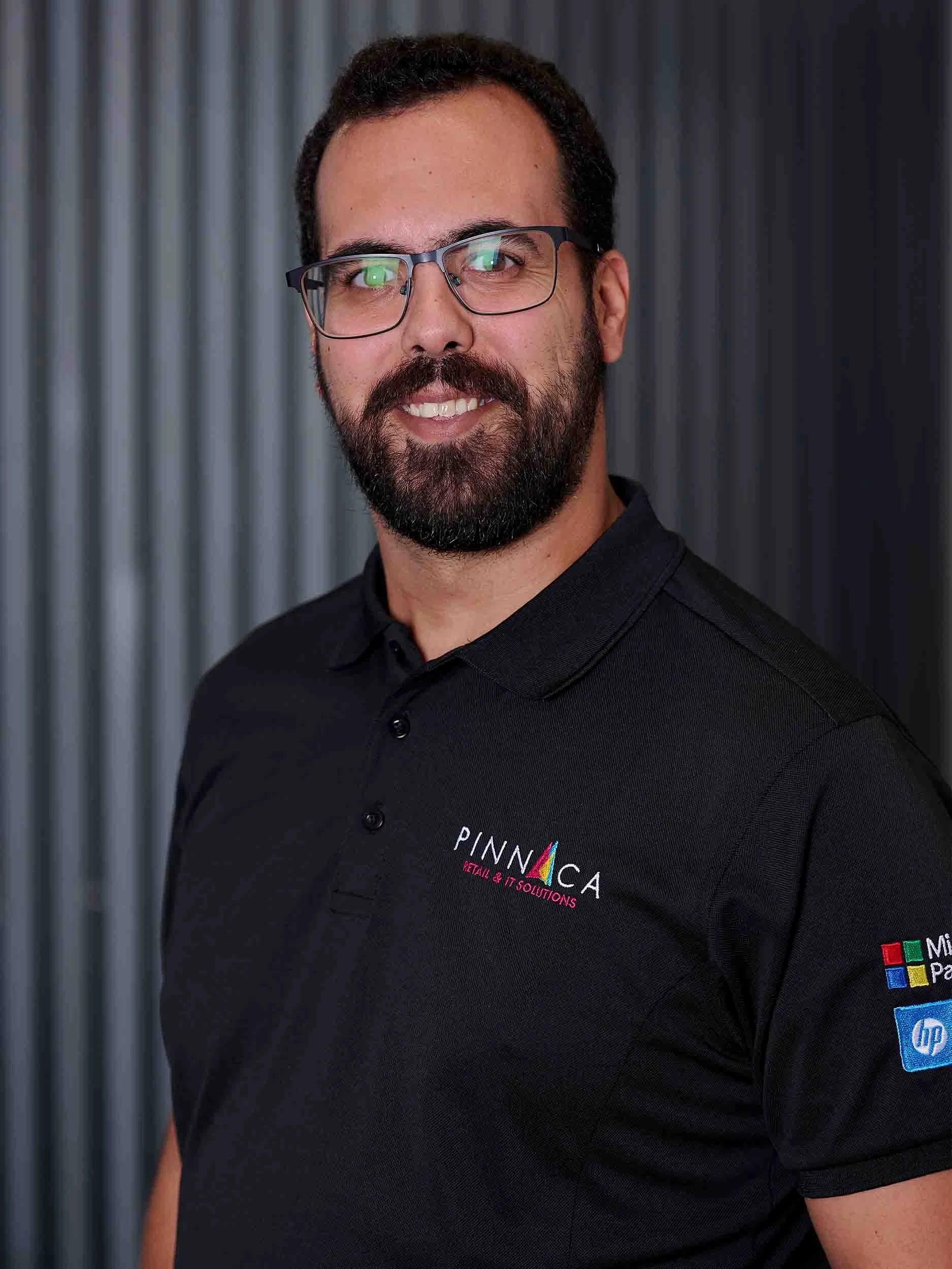 A man with glasses, a beard, and dark hair, wearing a black polo shirt with logos for Pinnaca and Microsoft, standing in front of a gray metallic background.