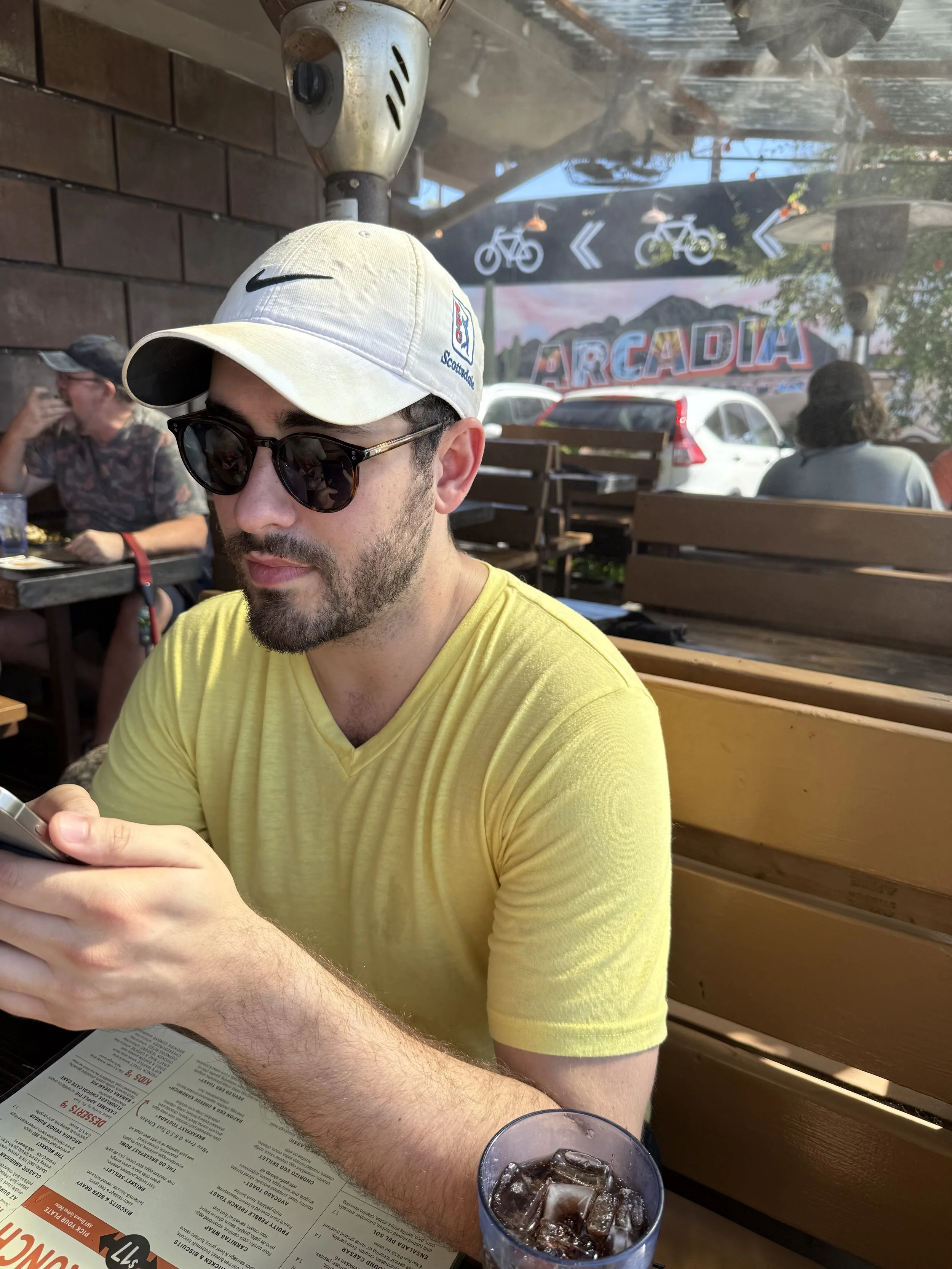 A man with a beard wearing sunglasses, a yellow T-shirt, and a white cap, sitting at an outdoor restaurant table, looking at his phone. There is a menu on the table and a glass of dark soda with ice in front of him. In the background, there are other