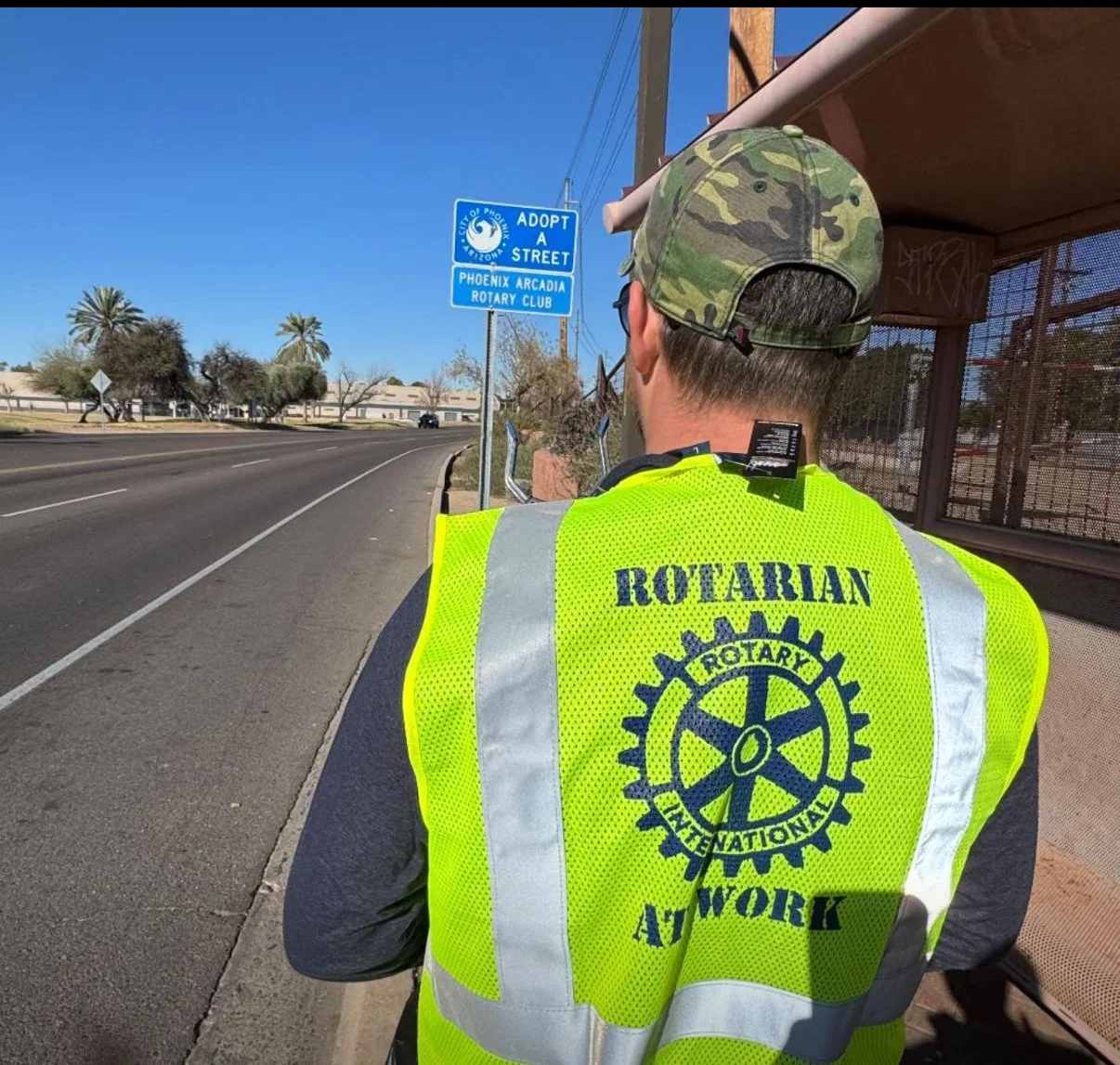 Person in a yellow high-visibility vest with the Rotary International logo and the words 'Rotarian At Work' printed on the back, standing on a sidewalk near a bus stop. The person is wearing a camouflage cap and glasses, facing away from the camera, 