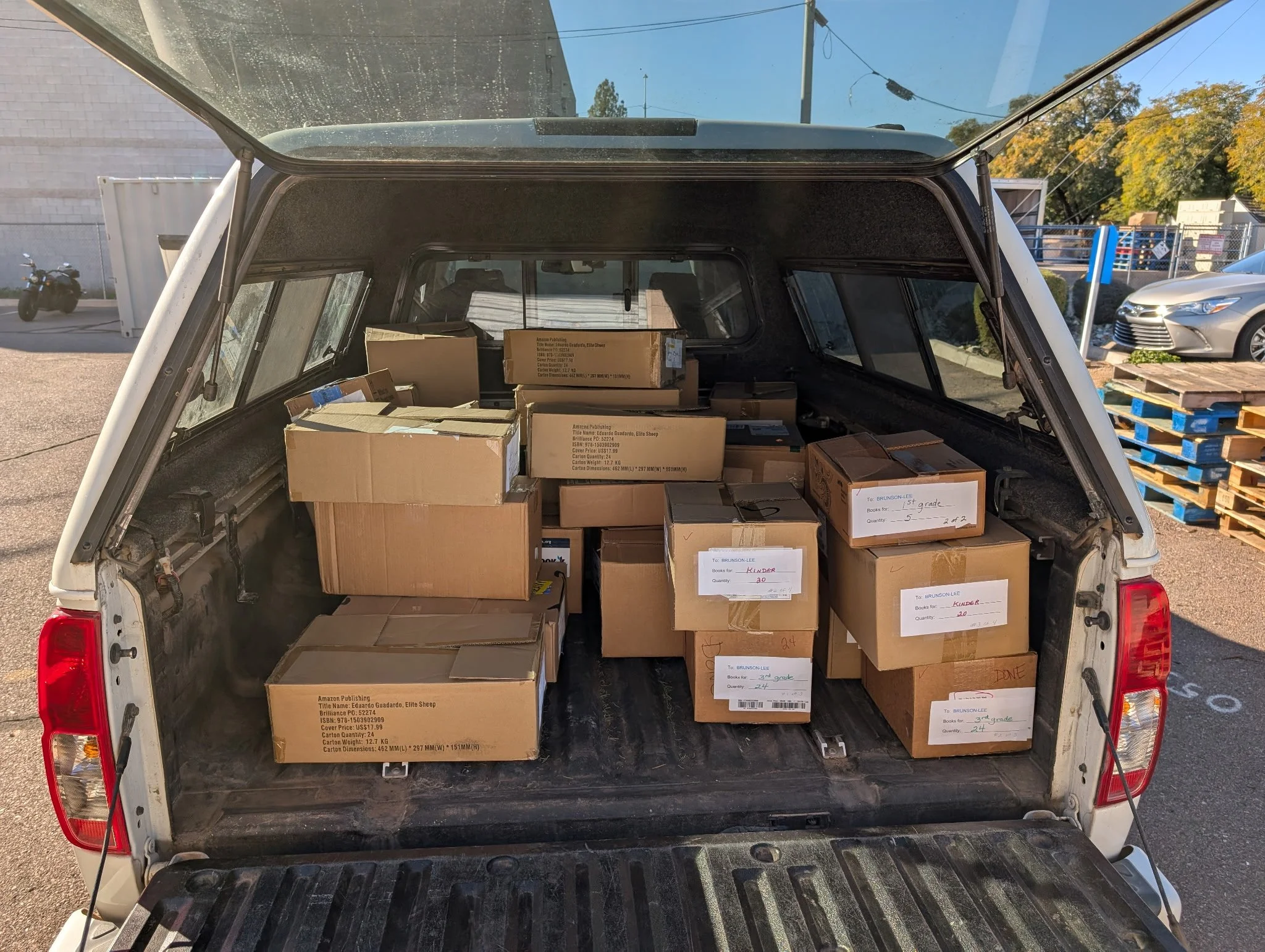 The back of a pickup truck filled with packed cardboard boxes, parked outdoors in a lot with a few other vehicles and pallets.