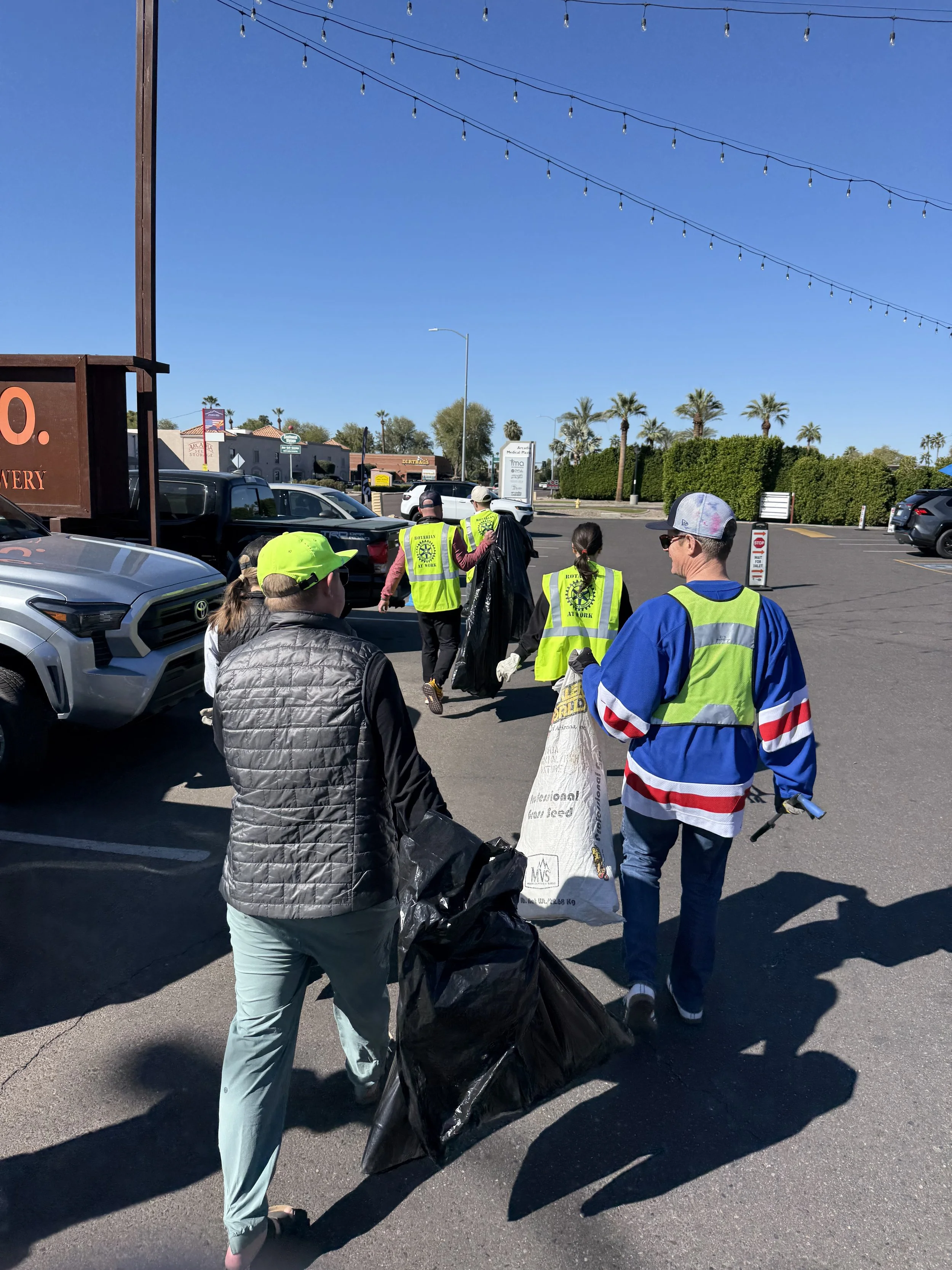 Group of people participating in a community cleanup in a parking lot, wearing yellow safety vests and carrying trash bags.