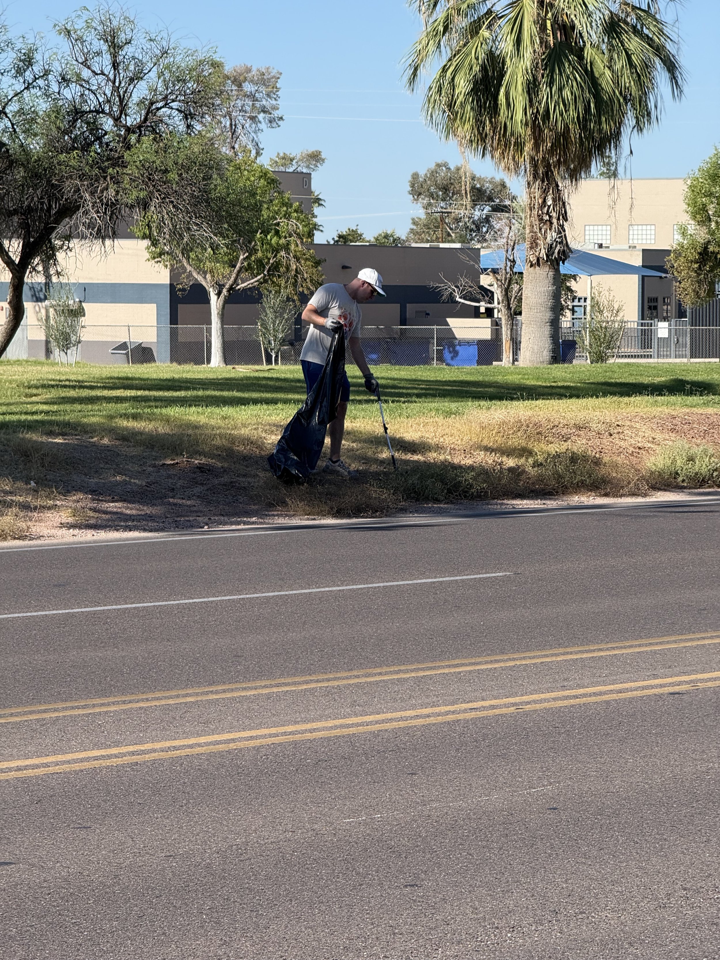 Person cleaning up trash along the roadside in a park with trees and buildings in the background.