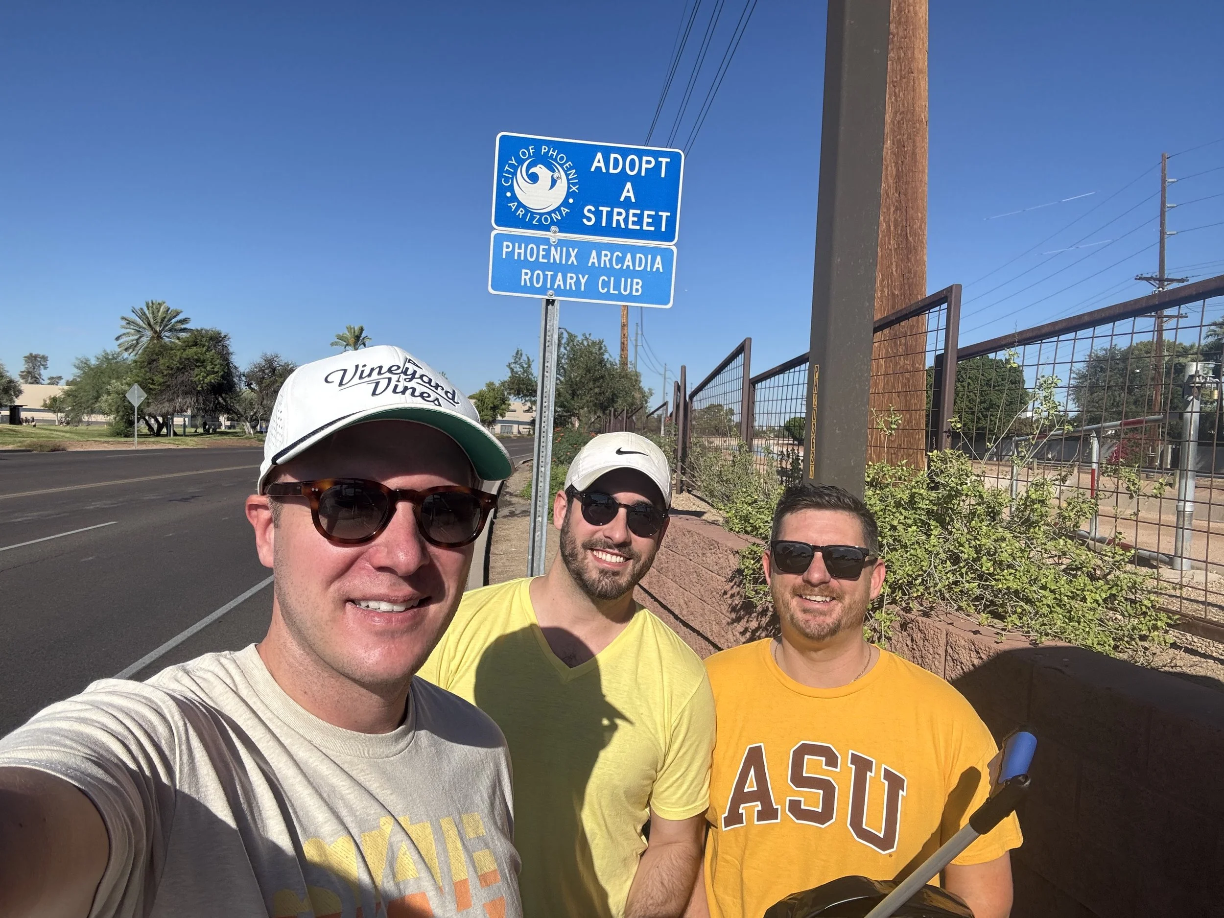 Three men taking a selfie outdoors on a sunny day near a street in Phoenix, Arizona, with a blue sign that says 'Adopt a Street' and 'Phoenix Arcadia Rotary Club' in the background.
