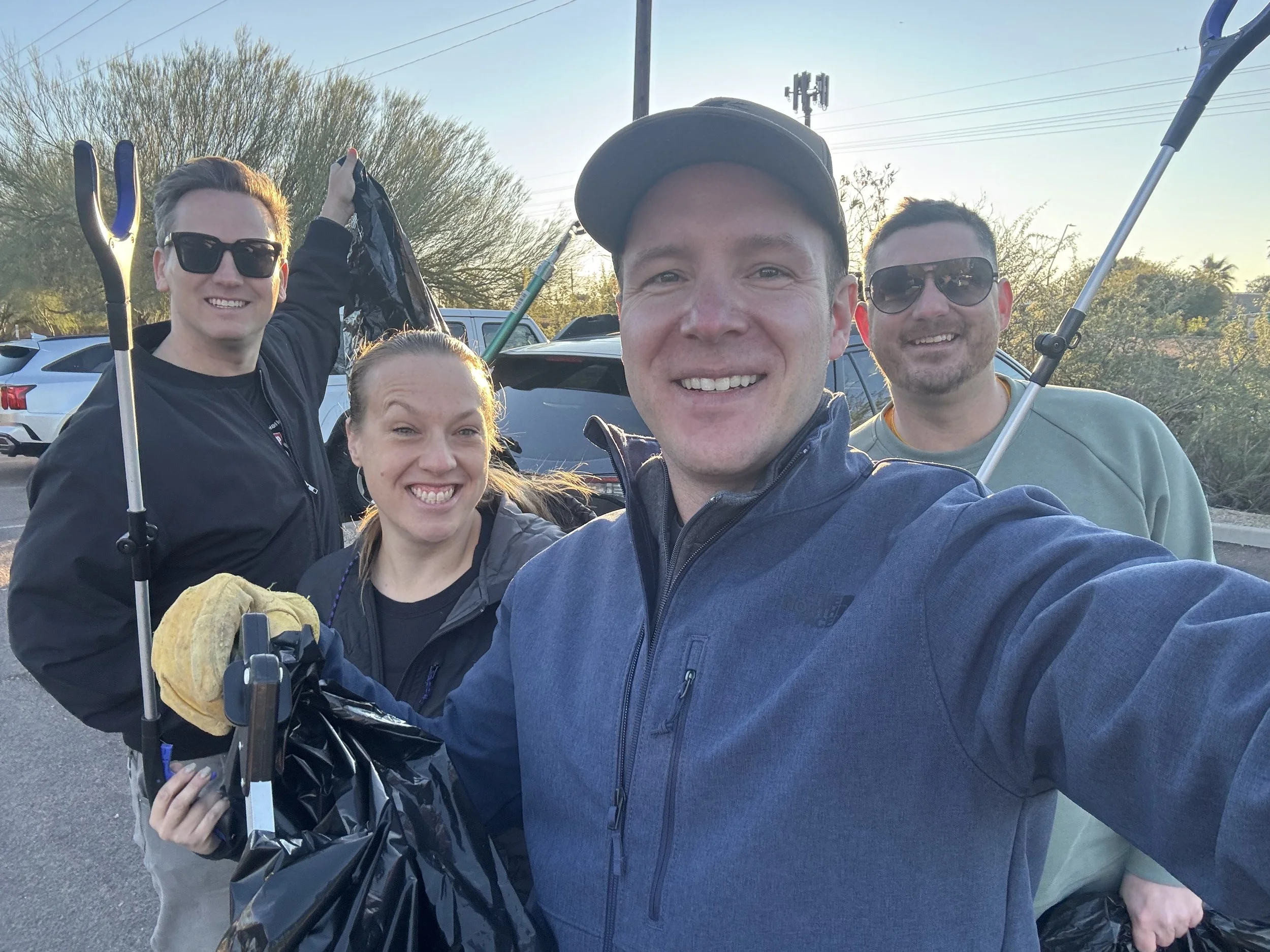 Four people smiling outdoors during what appears to be a clean-up event, holding trash pick-up tools and a trash bag. The background shows parked cars and trees at sunset.