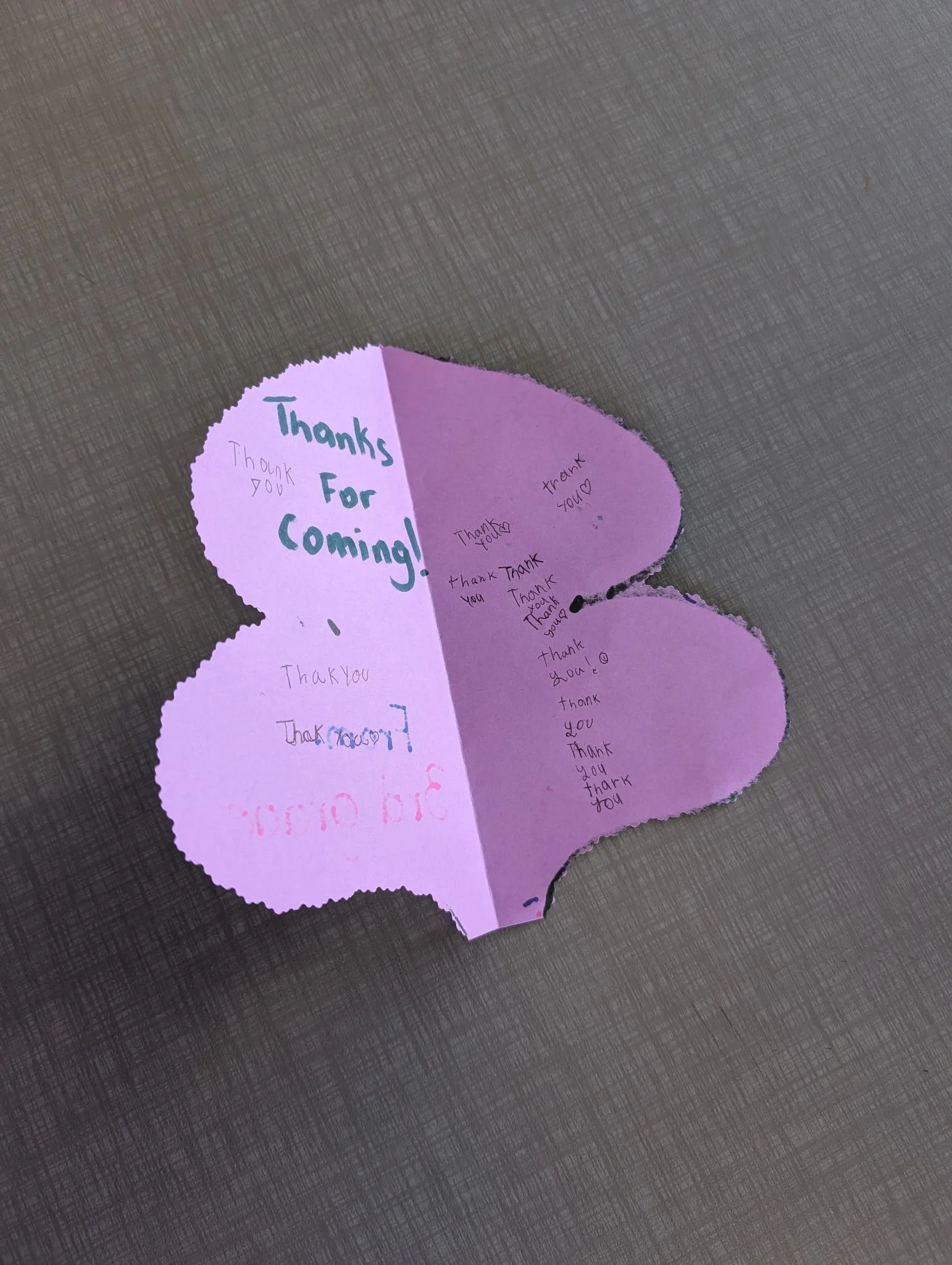 Purple paper cutout shaped like a heart with various handwritten 'Thank you' messages, placed on a textured surface.