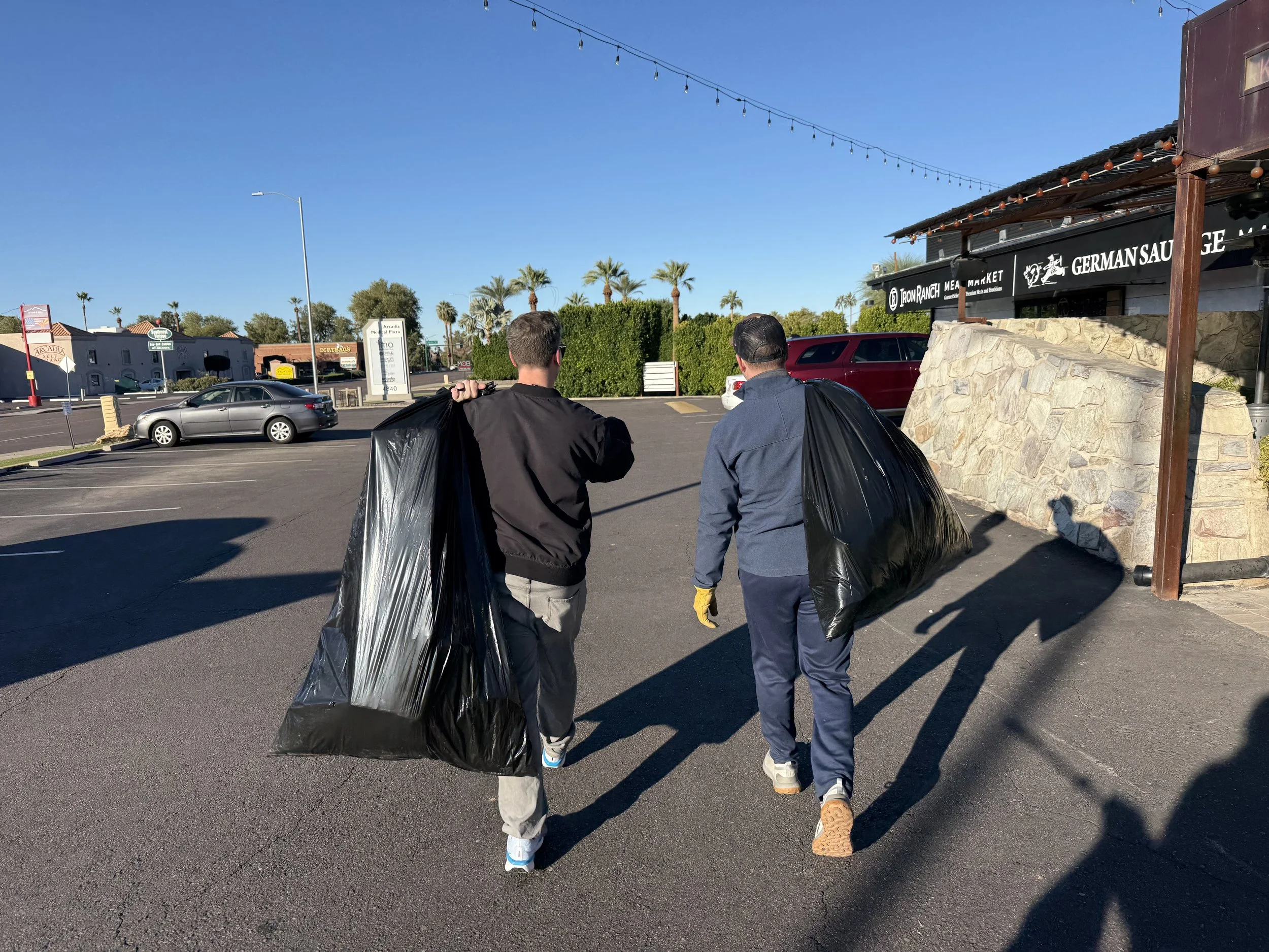 Two men walking away on a parking lot while carrying large black plastic trash bags, with a restaurant and cars in the background under a clear blue sky.