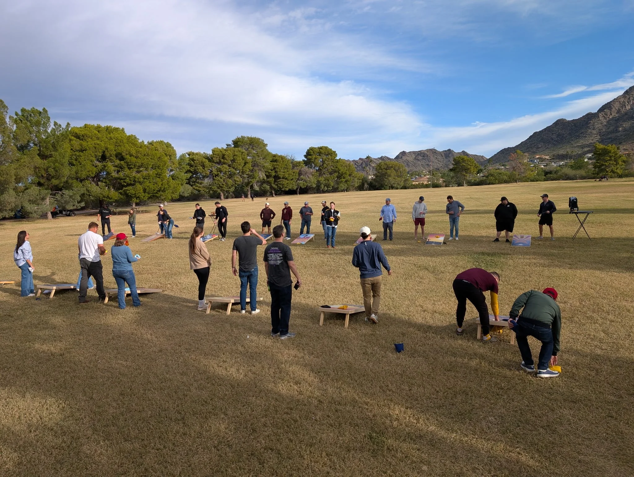 A large outdoor gathering with people playing cornhole on a grassy field, with green trees, mountains, and a partly cloudy sky in the background.