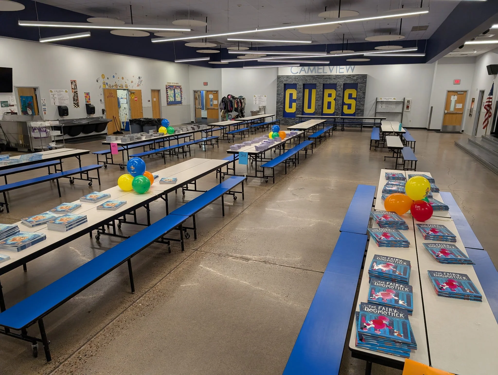 Empty cafeteria with long tables decorated with balloons and stacks of books,, and a wall with 'CUBS' letters in the background.