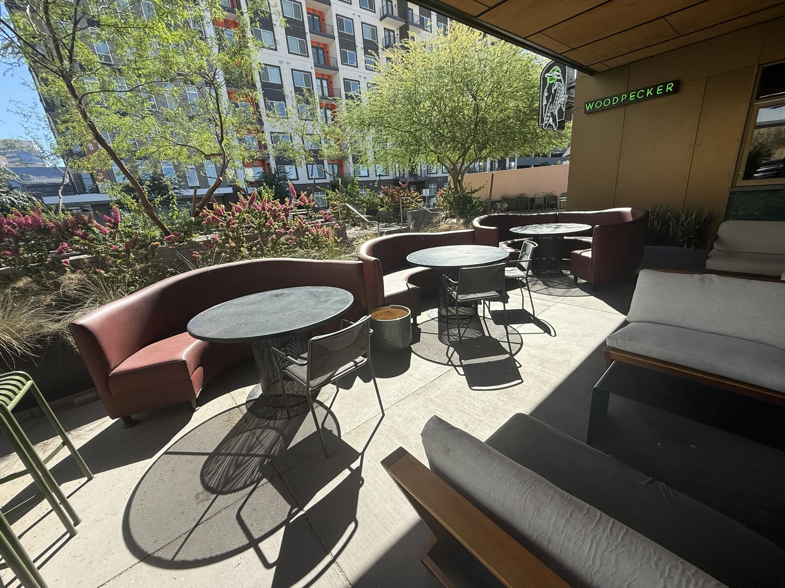 Park Centrals best bar 'The Green Woodpecker' Location of Arcadia Rotary clubs Annual Poker Tournament. An outdoor patio area with round tables, curved booths, and lounge chairs, surrounded by plants and trees, with a tall buildings.
