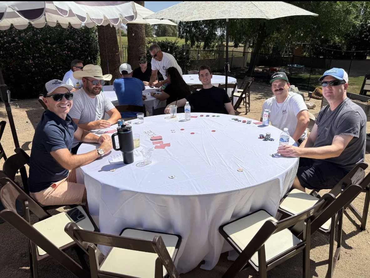 Group of people sitting around a large round table outdoors under large umbrellas, playing a card game on a sunny day.