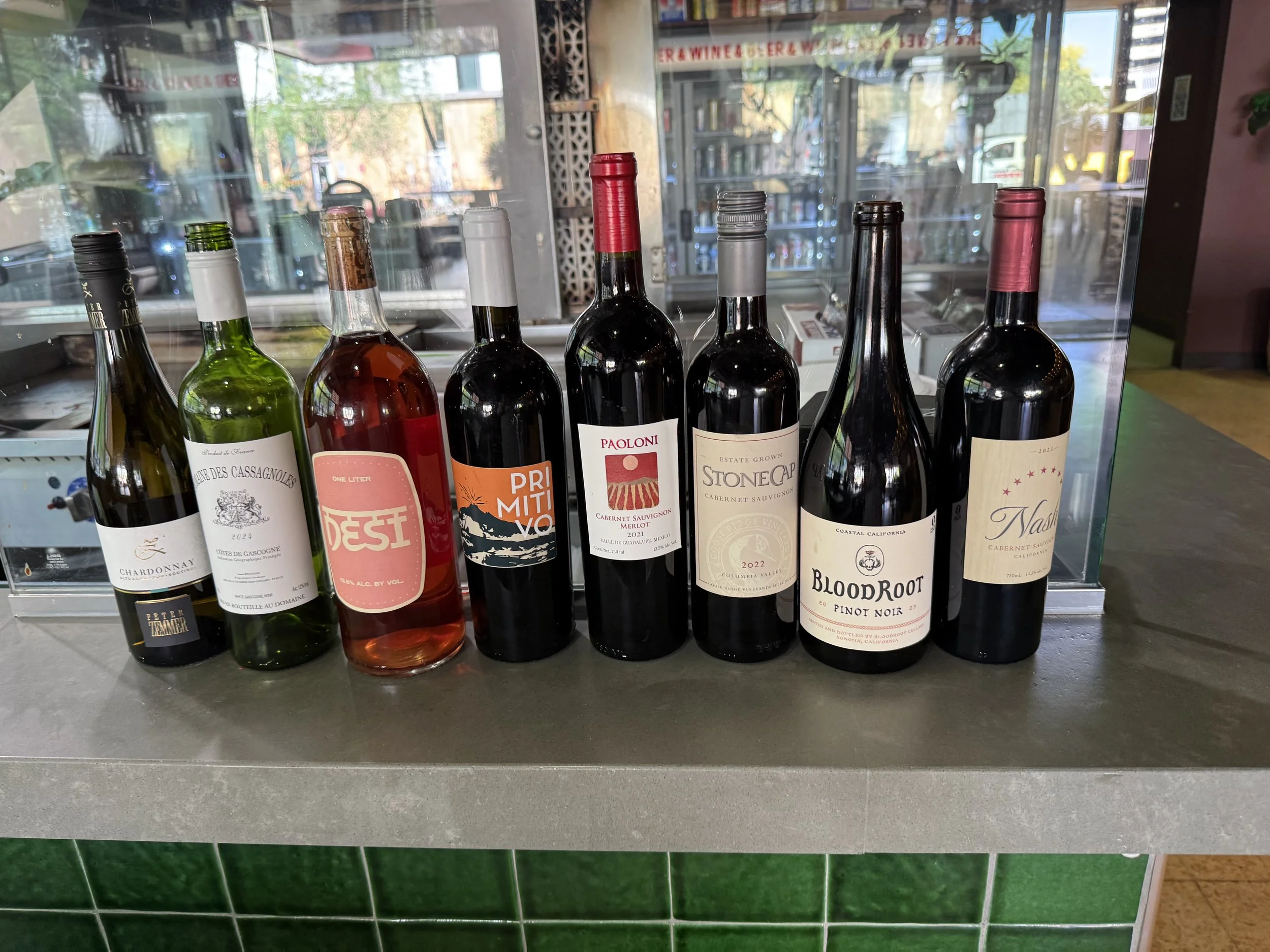 Seven bottles of wine lined up on a counter in front of a glass window, with a view of the street outside.