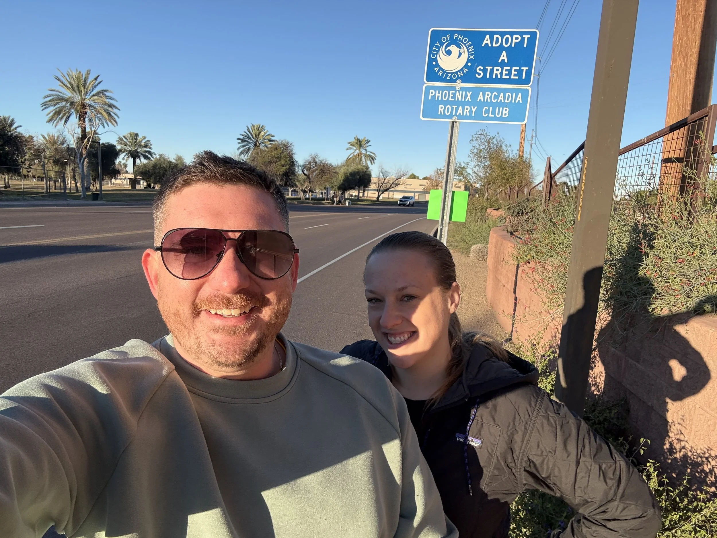 A man and woman taking a selfie on a sidewalk in Phoenix, Arizona, with palm trees and a blue street sign that reads 'Adopt a Street', 'City of Phoenix', 'Phoenix Arcadia Rotary Club' in the background.