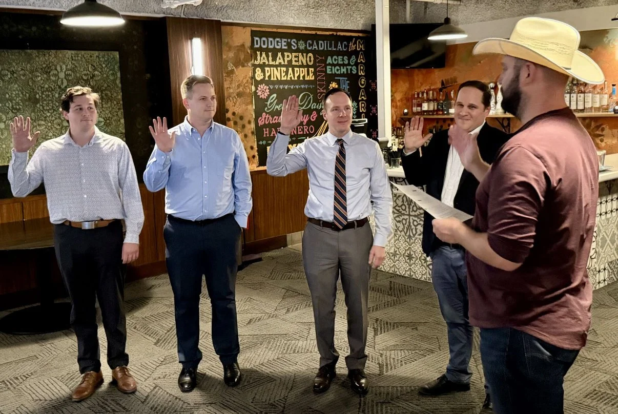 Five men standing in a room, raising their right hands for a formal oath or pledge, with one man wearing a yellow cowboy hat reading from a paper. Swearing in our 2025-2026 Rotarian Board at our Annual Rotarian Dinner.