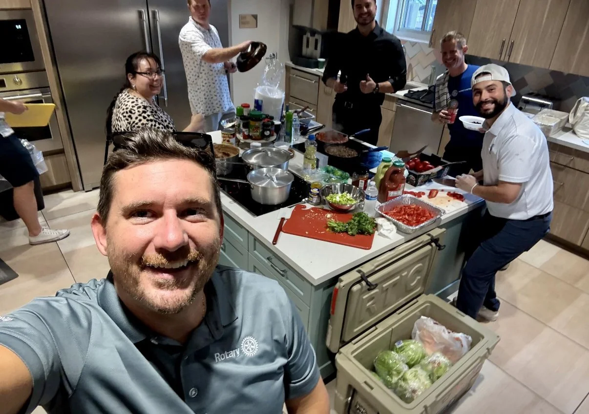 Group of people cooking and preparing food in a kitchen, smiling and looking at the camera, with various ingredients and kitchen utensils on the countertop. At a Ronald McDonald House Charities event.