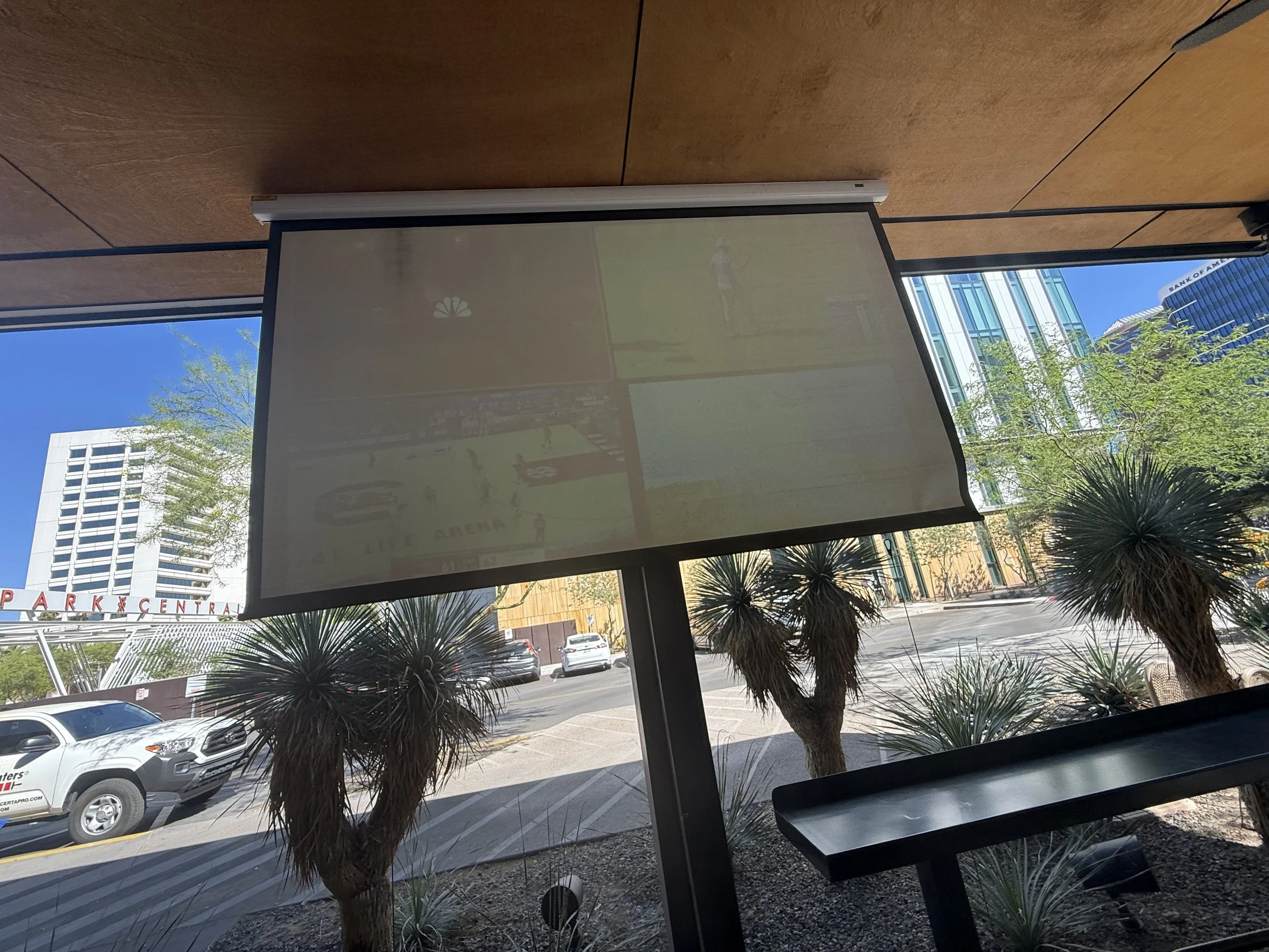 East Facing Shaded seating area overlooking a street through large windows with trees, parked cars, and modern buildings in the background, with a projection screen  to watch sports on the Bar patio for the PARC Poker Tournament 