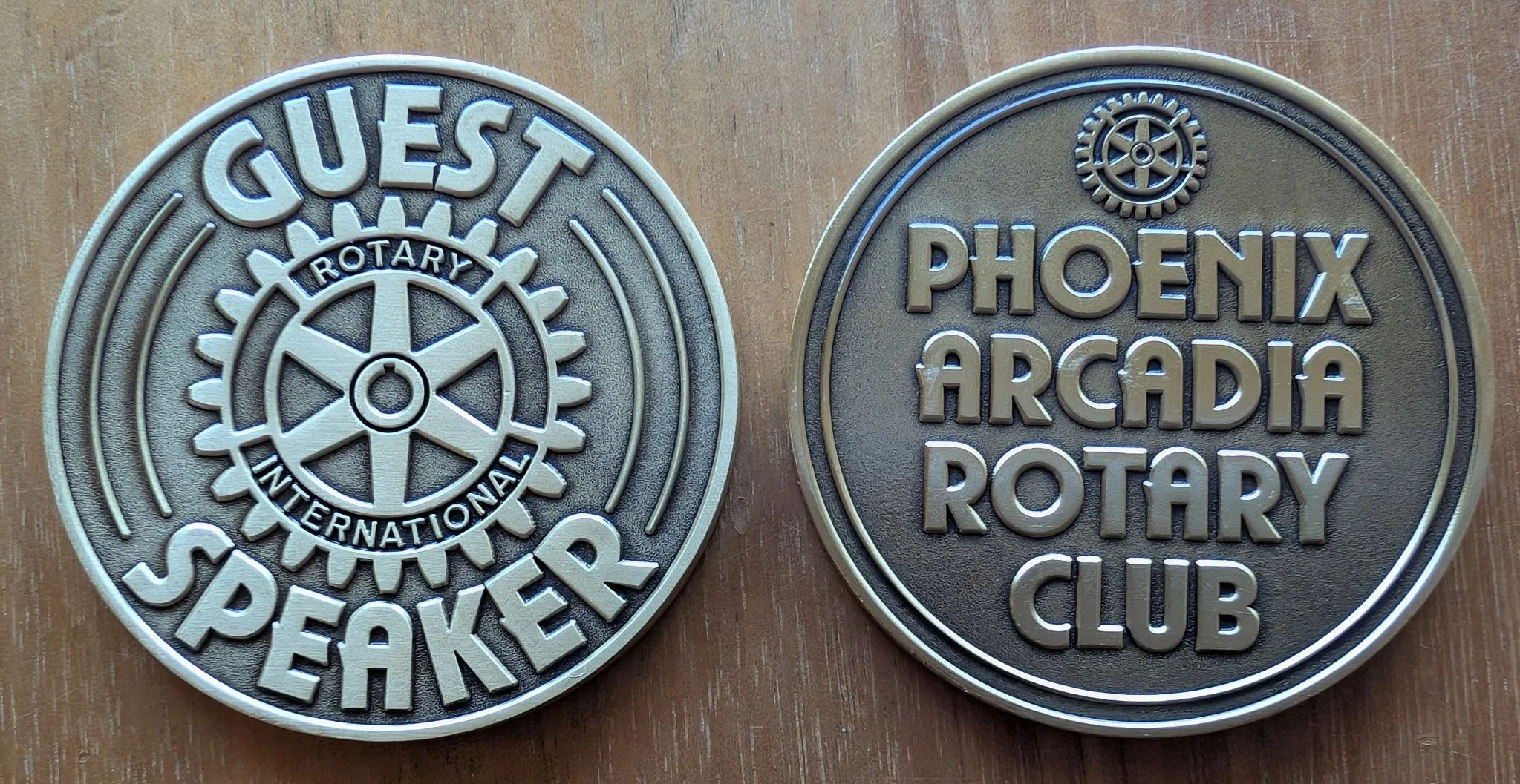 Two circular metal coins, one side  reads "QUIET SPEAKER" with a rotary gear wheel in the center and the words "ROTARY INTERNATIONAL," the other reads "PHOENIX ARCADIA ROTARY CLUB" with a gear wheel emblem at the top. Coin Given to Guest Speakers