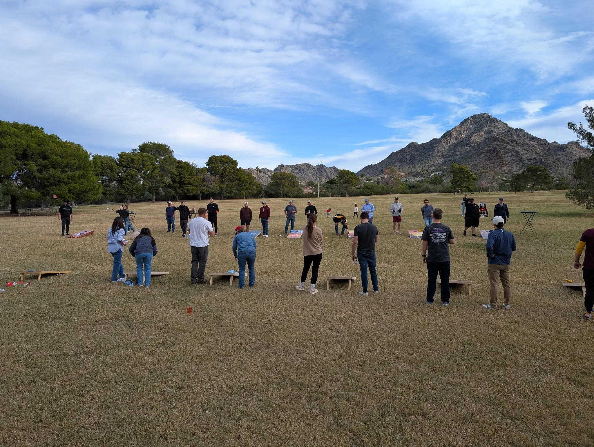 A group of people playing cornhole outdoors in a large grassy field with mountains and trees in the background under a partly cloudy sky.