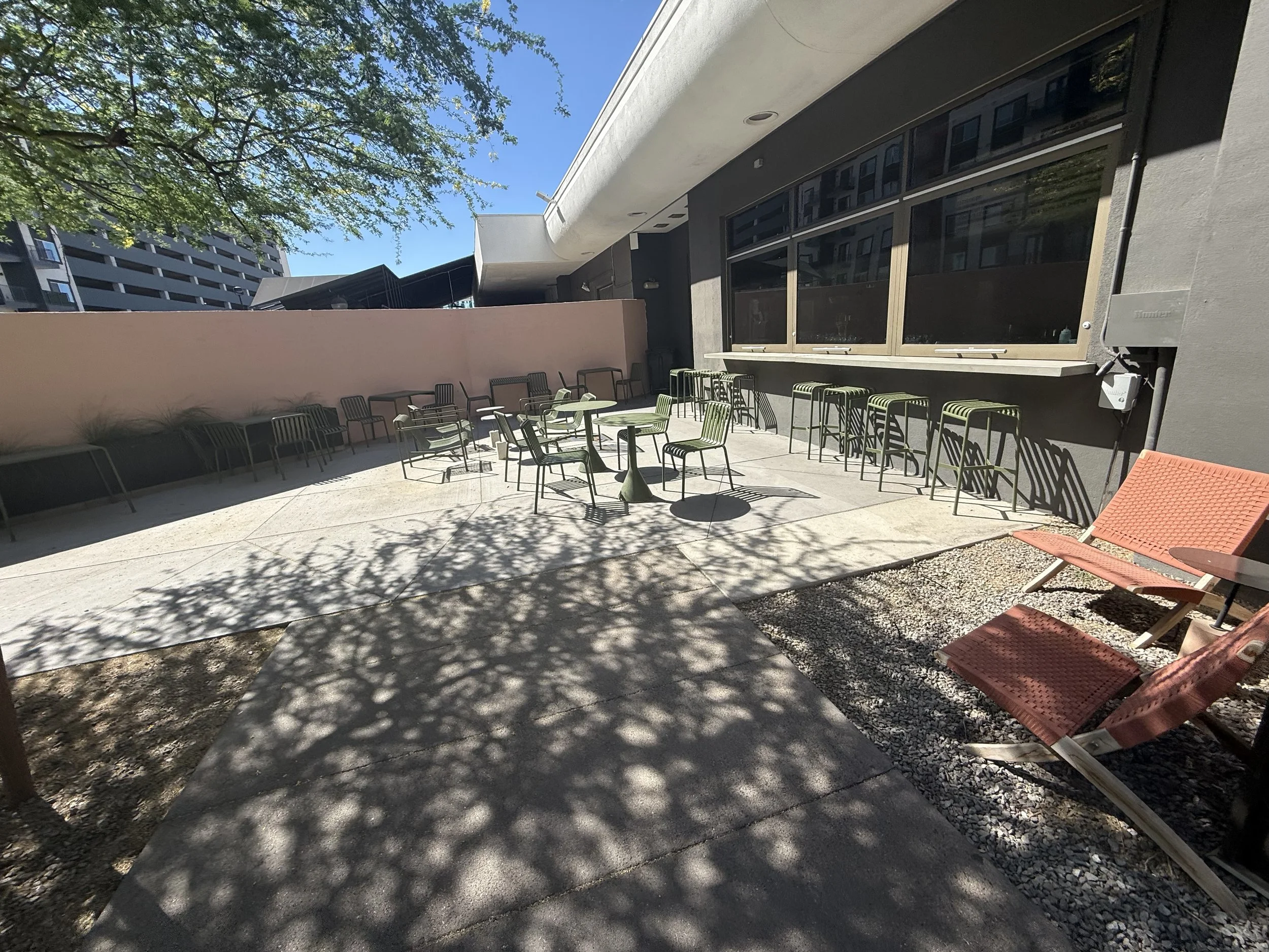 Outdoor patio with chairs and tables, some chairs are stacked, a pink wall, a gray building wall with large windows, and shadows of leaves on the ground and wall, in a sunny setting. Location for PARC annual poker tournament at the green woodpecker