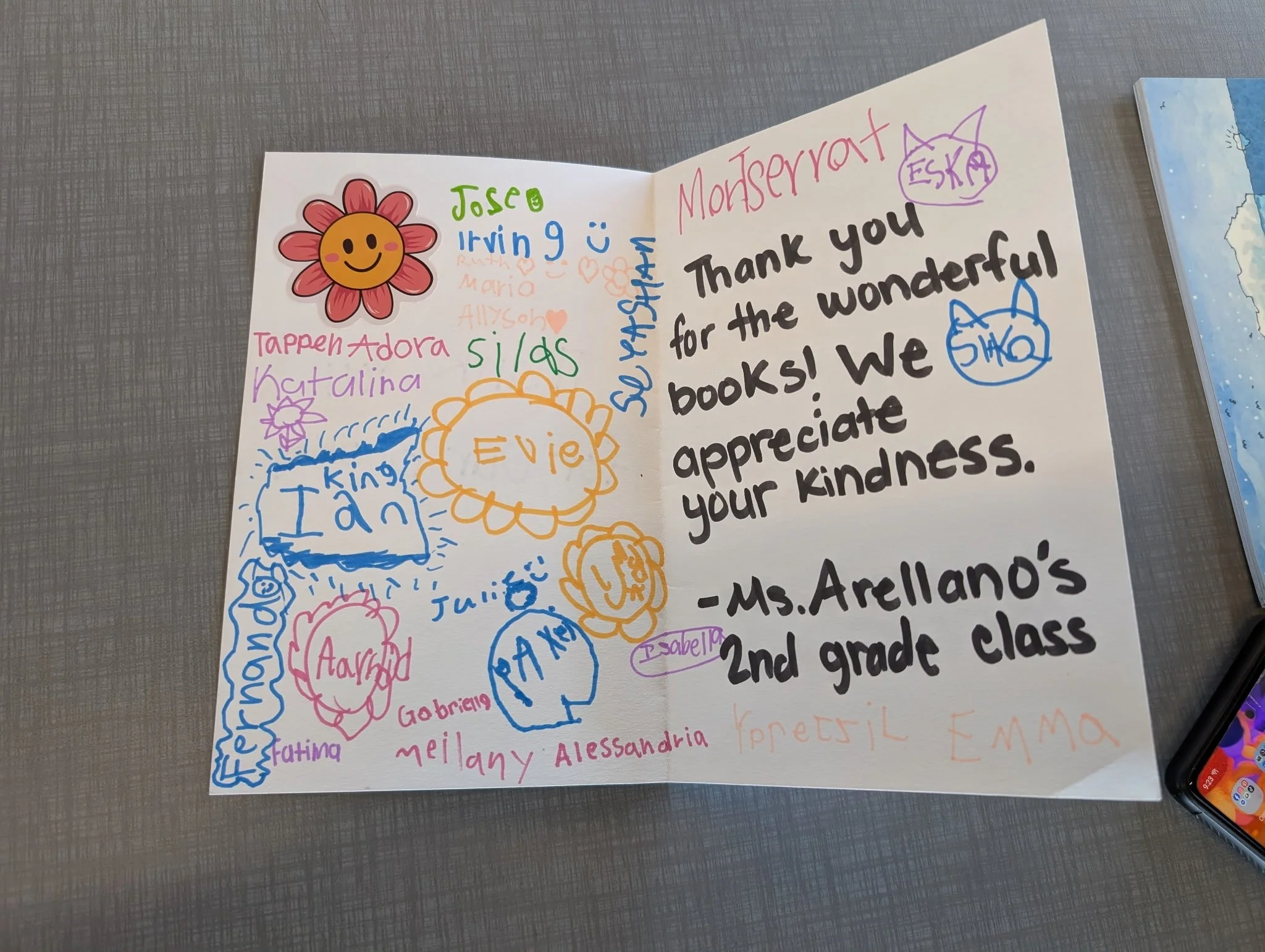 A handwritten thank you note for a library, with colorful drawings and signatures from second-grade students. The note includes a large message in black ink, a flower doodle, and several smaller names and doodles.
